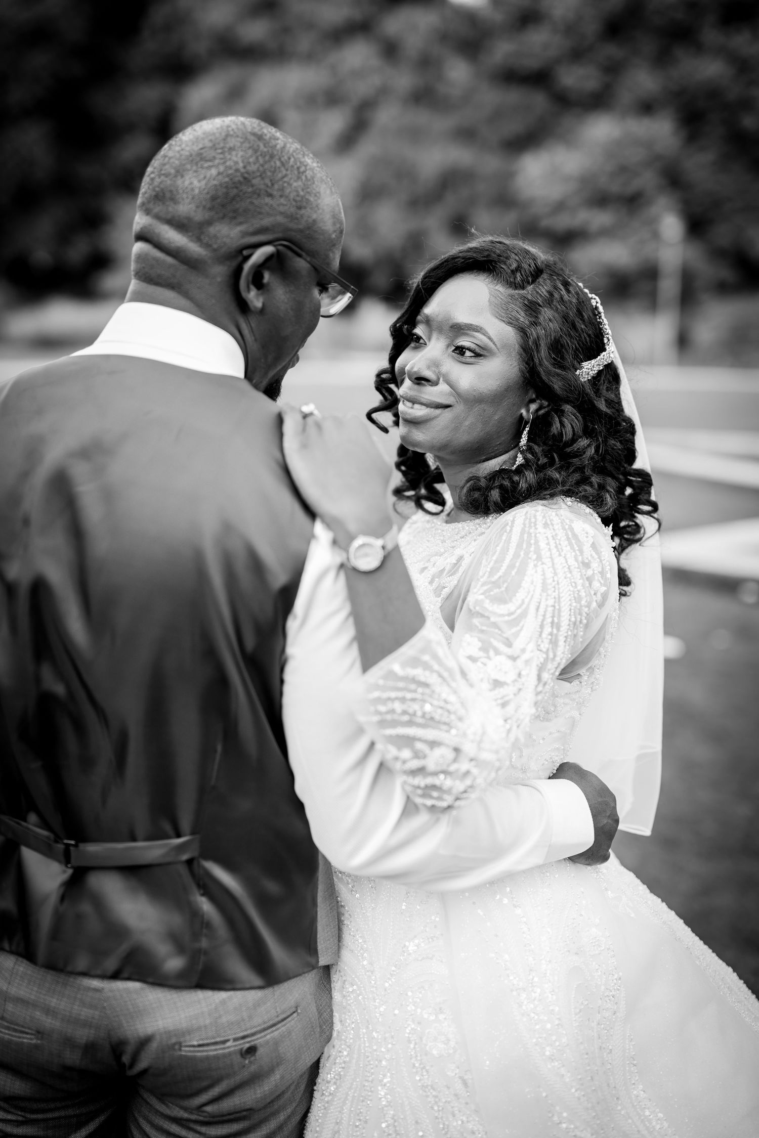 Black and white wedding photo of a bride in a beaded gown smiling at her groom during their first dance outdoors.