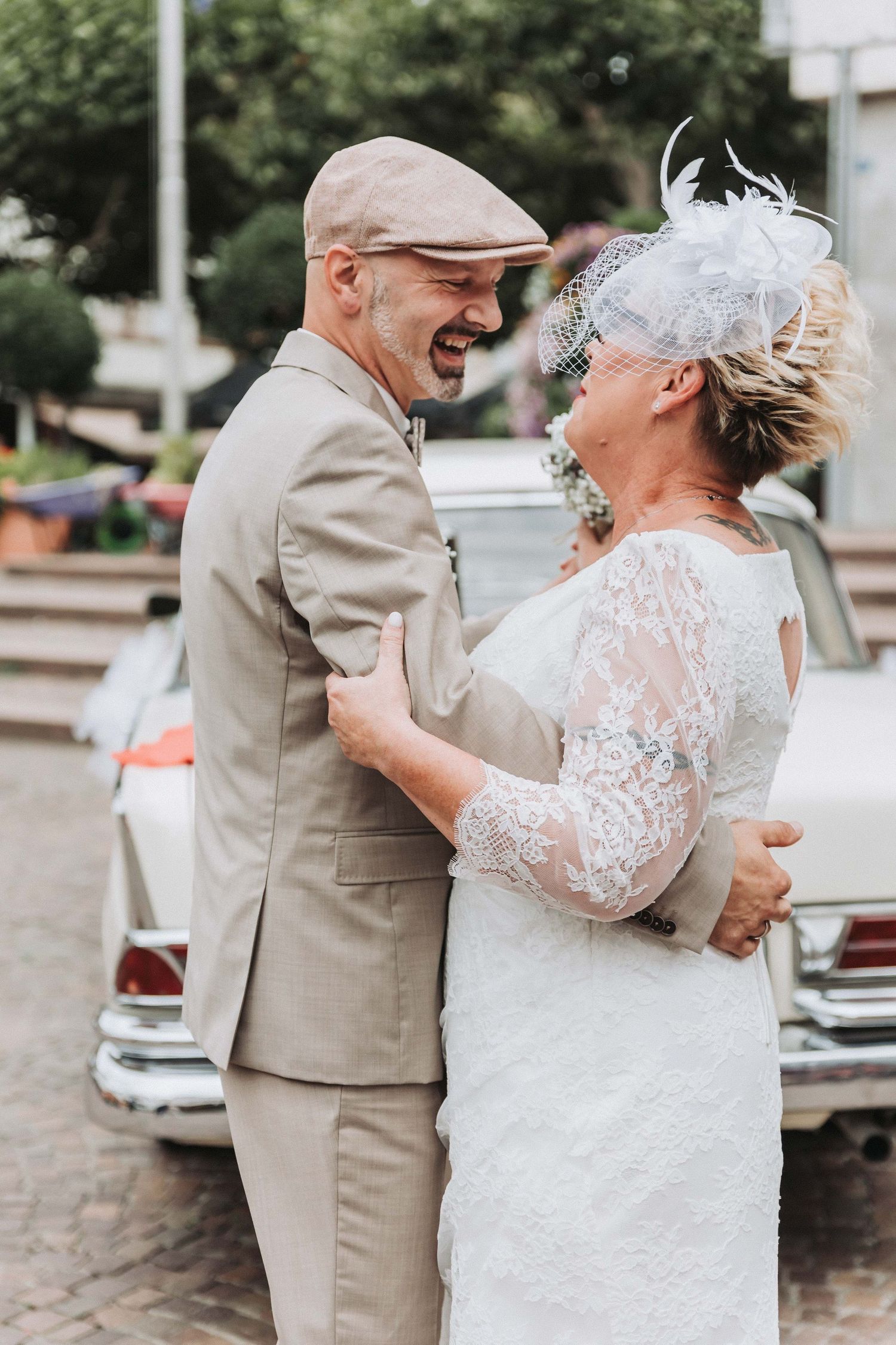 A couple shares a joyful moment together in formal attire, with the person in a white lace dress and tan vest embracing.