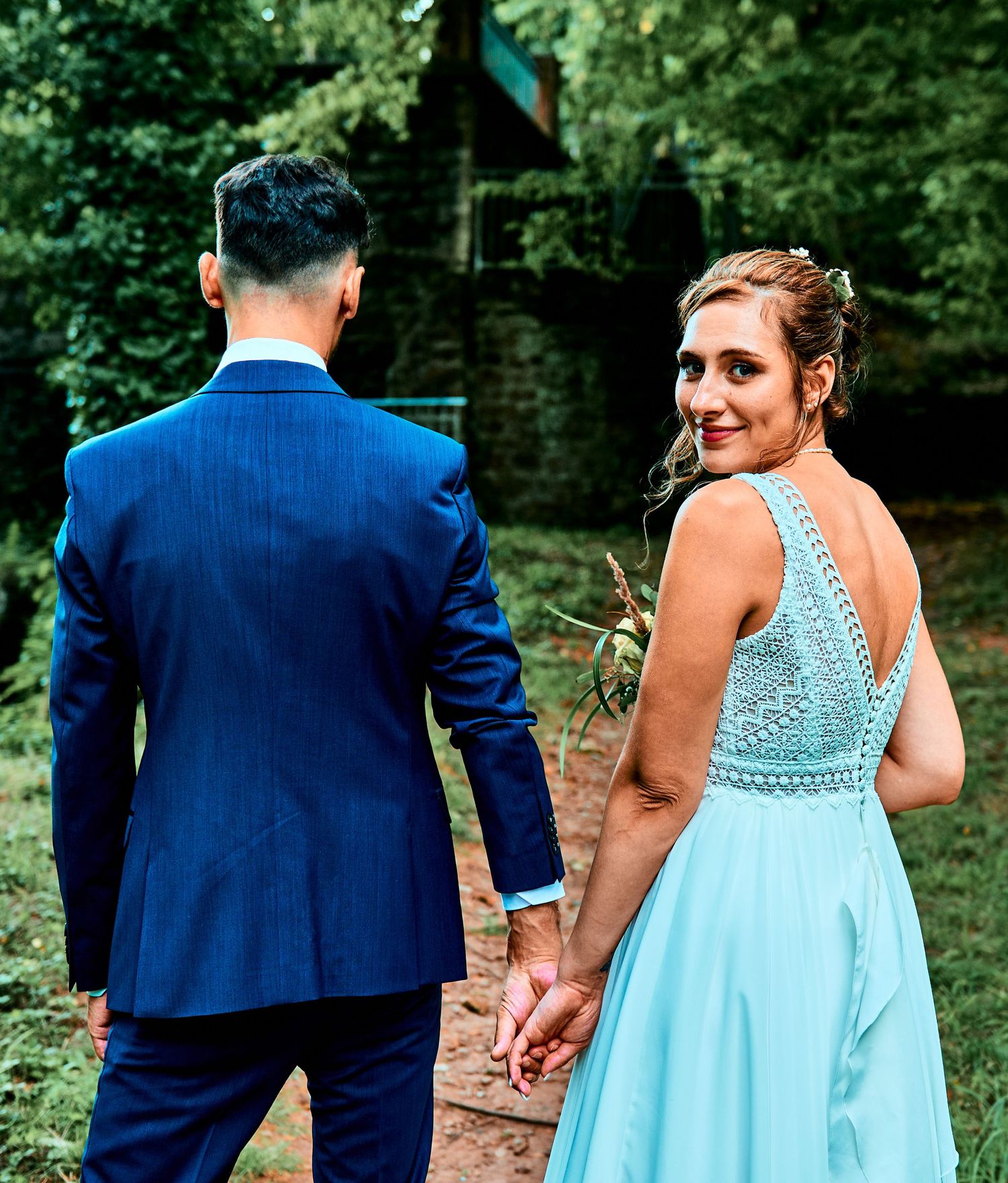 A couple in formal attire holding hands while walking outdoors, with the woman wearing a light blue gown and man in navy suit.