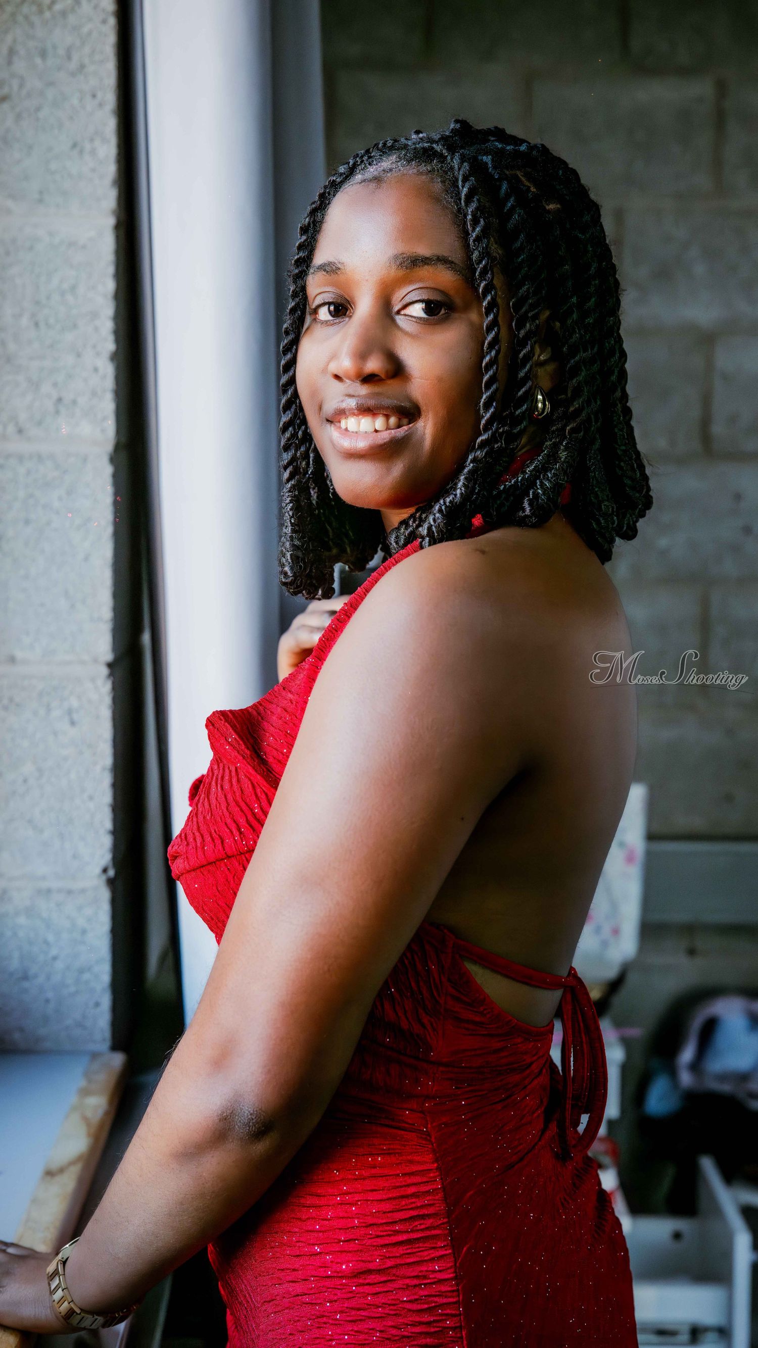 Portrait of someone in a vibrant red dress with braided hair smiling warmly by a window.