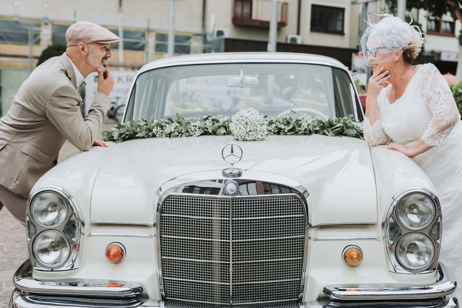 A classic silver Mercedes-Benz car decorated with white flowers and ribbons for a wedding celebration.