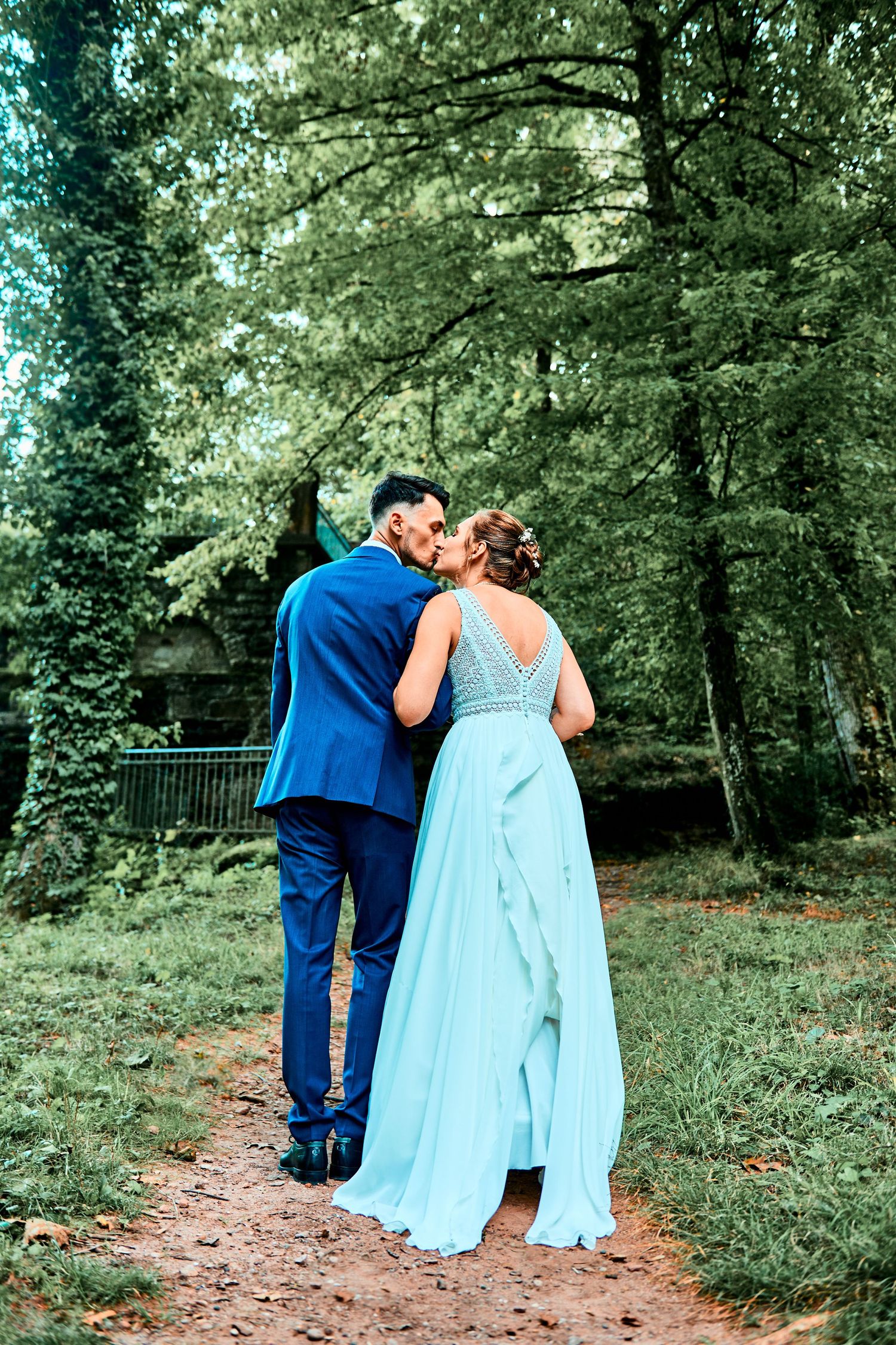 A couple in blue attire walking together on a forest path surrounded by lush green trees and foliage.