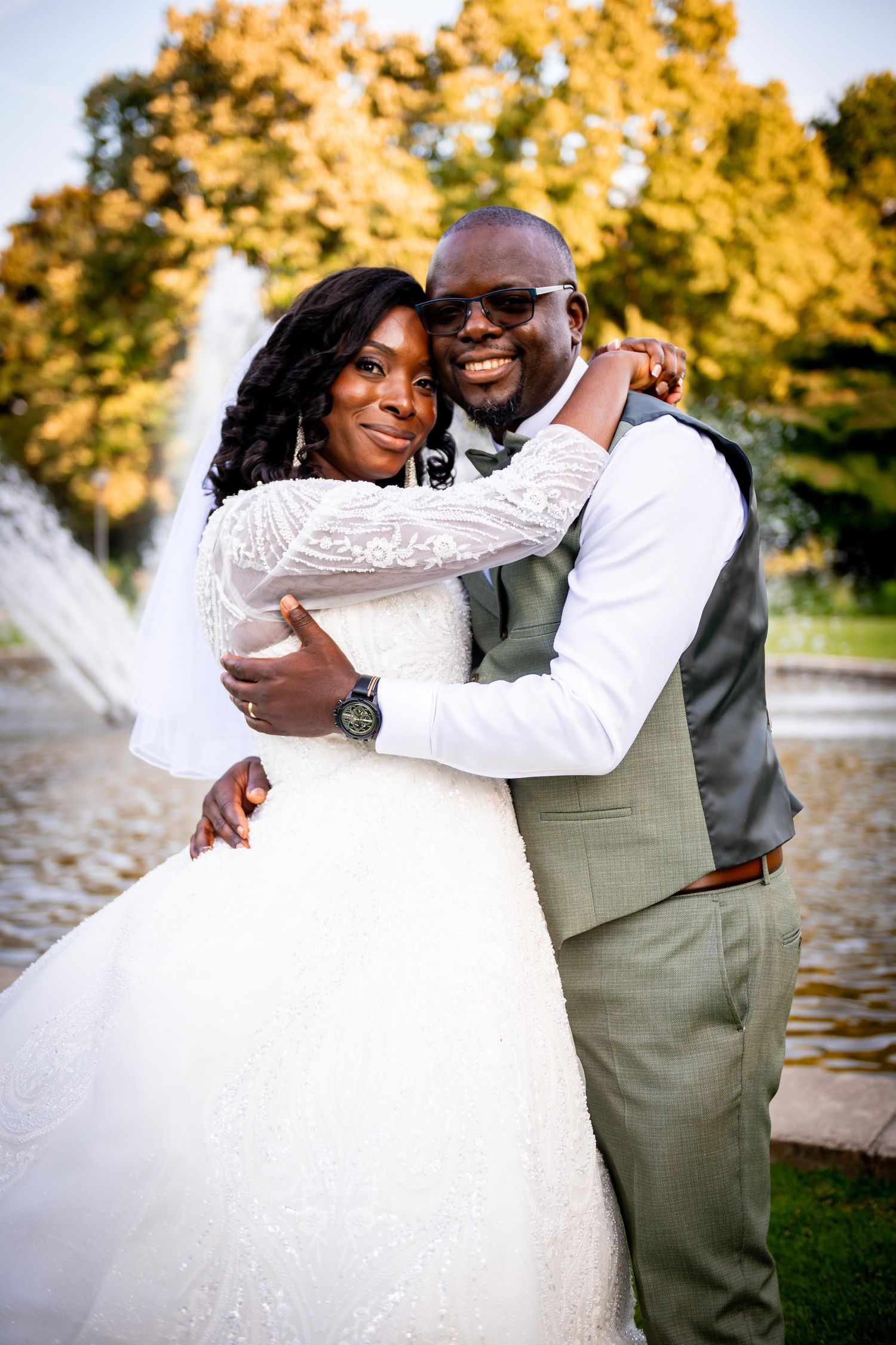 Bride and groom embrace smiling in front of a fountain, groom wearing gray suit and sunglasses.
