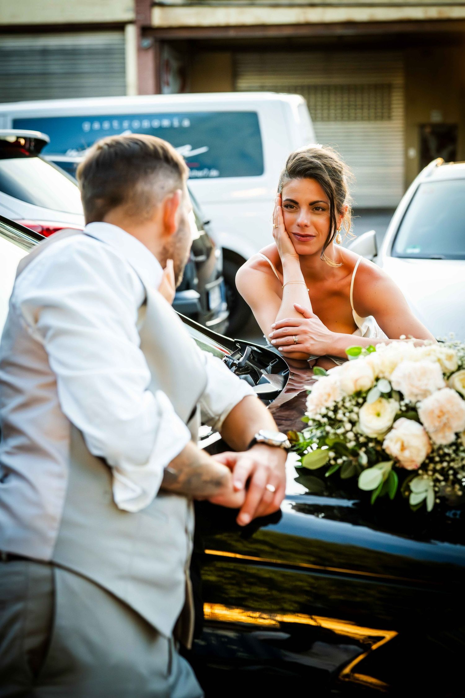 Bride in pink dress holds bouquet of white flowers beside black car, speaking with groom in white suit.