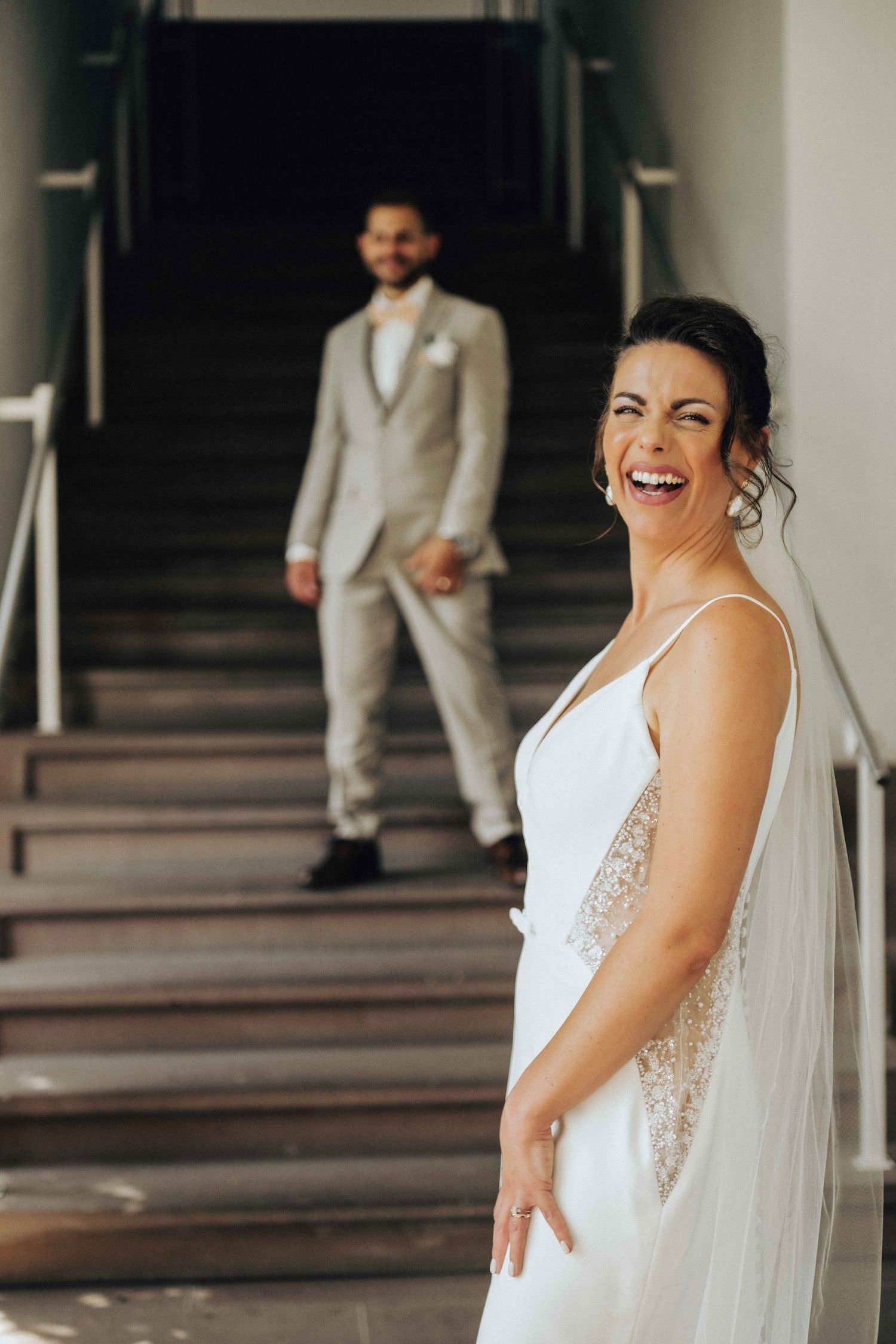 Smiling bride in white gown laughs near staircase as groom stands in background wearing a light gray suit.