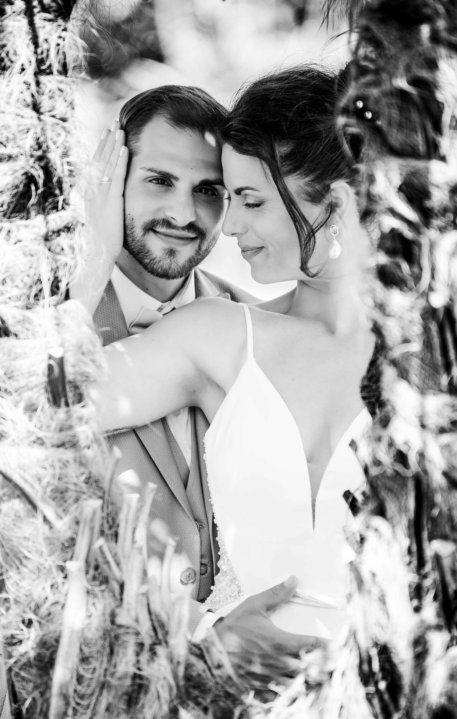 Black and white portrait of smiling groom embraced by bride in white dress among natural foliage.