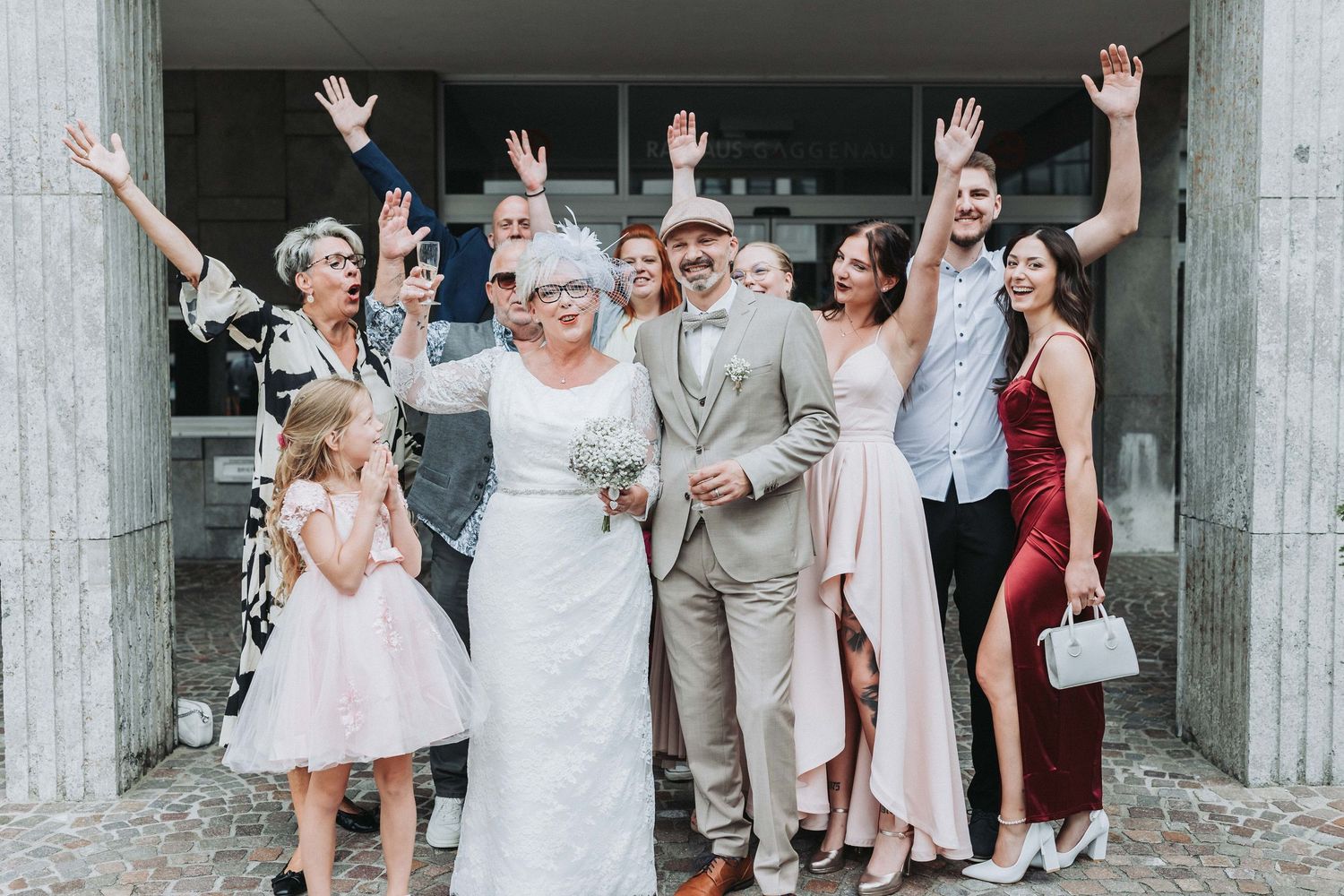 A joyful wedding party poses together in front of a concrete wall, celebrating with raised hands and smiles.