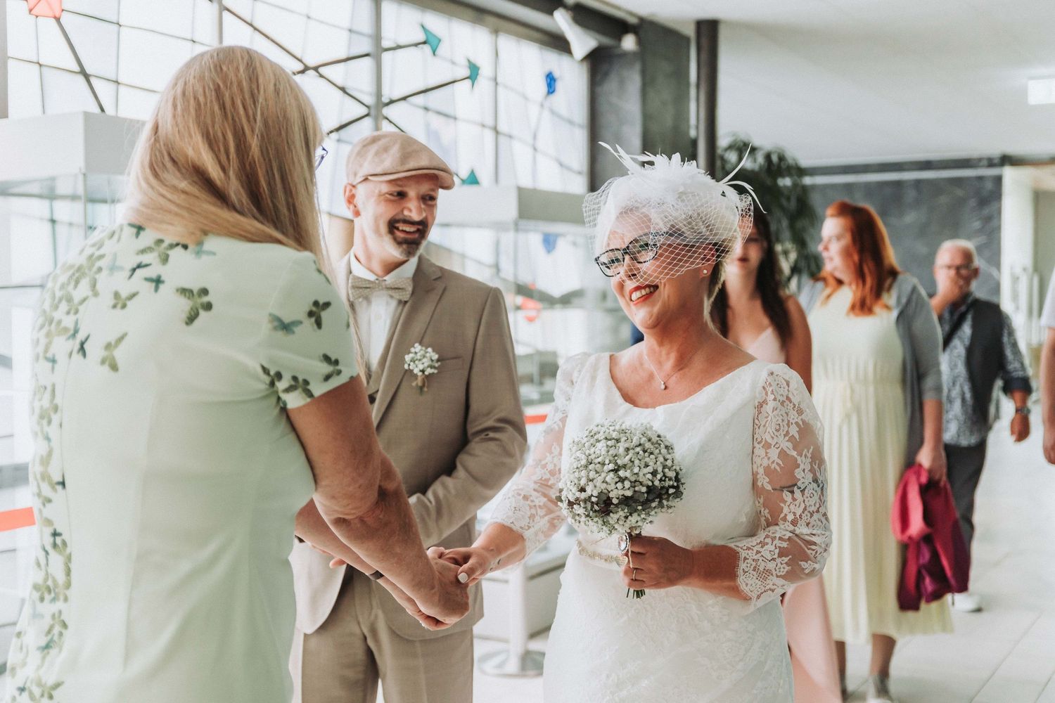 Wedding guests share joyful moments at an indoor reception venue with natural lighting and floral decor.