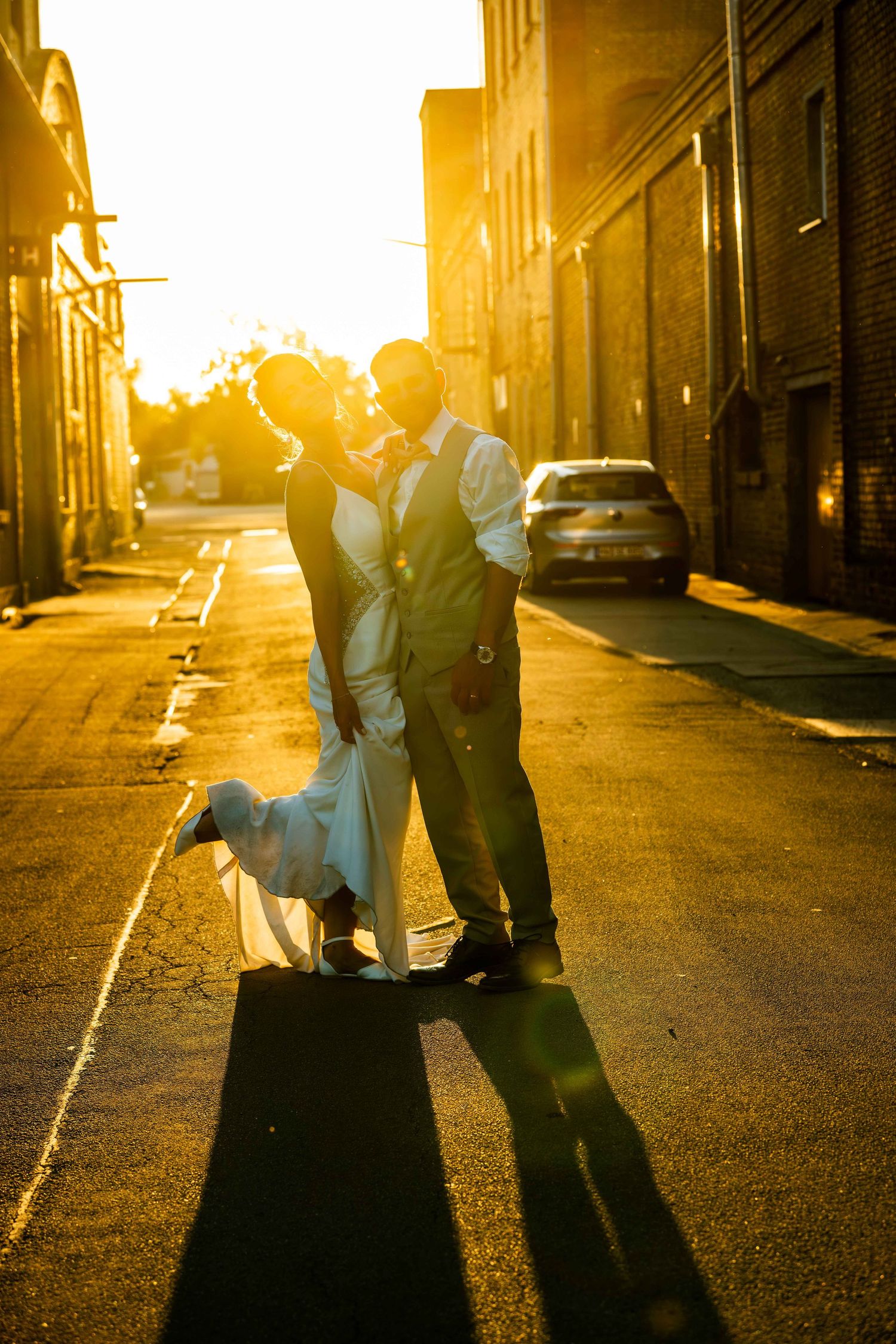 Bride and groom share a romantic kiss on a sunlit street during golden hour, casting long shadows on the pavement.