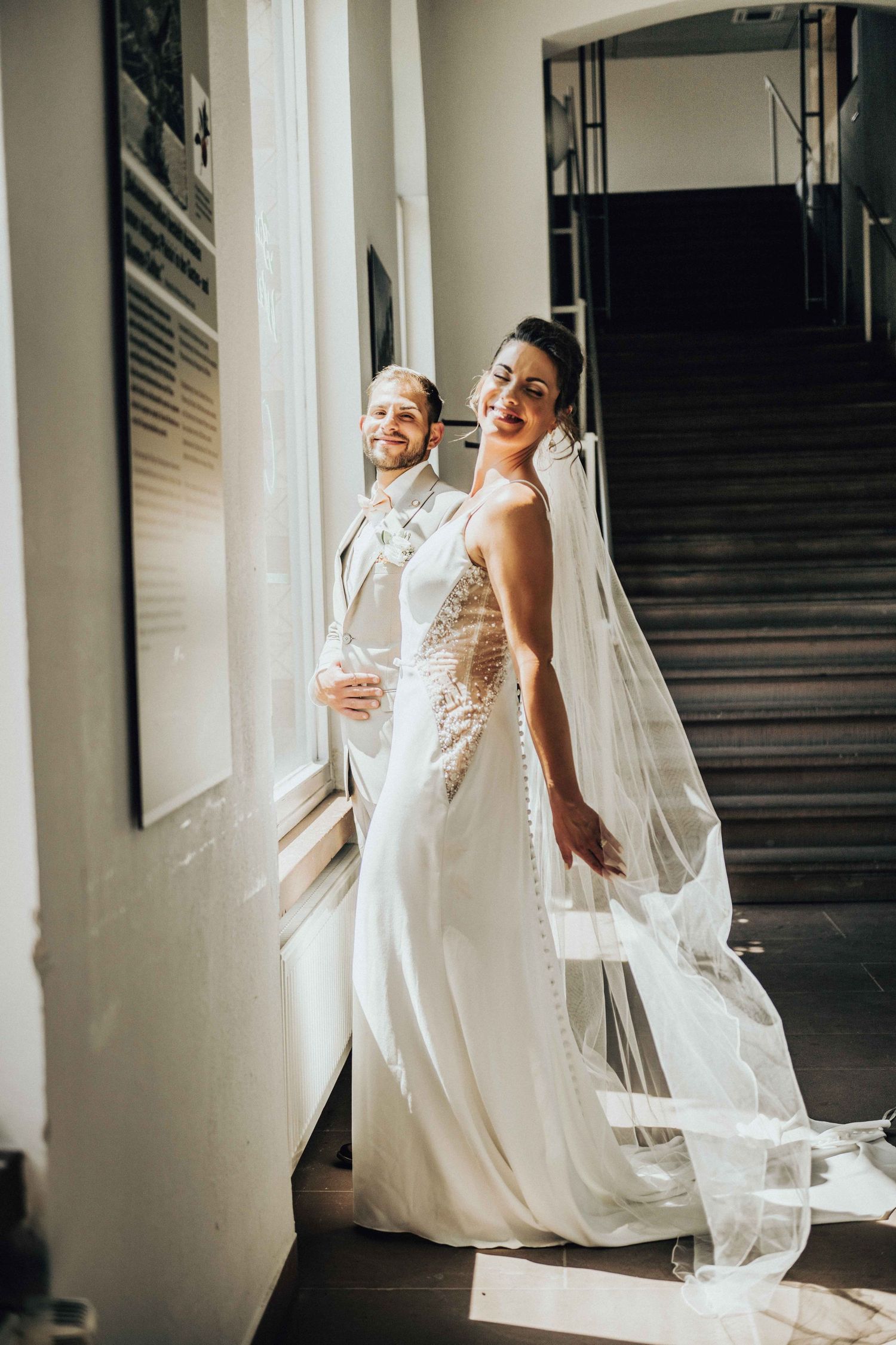 Smiling bride in white gown with flowing veil stands by window with groom in sunlit hallway.