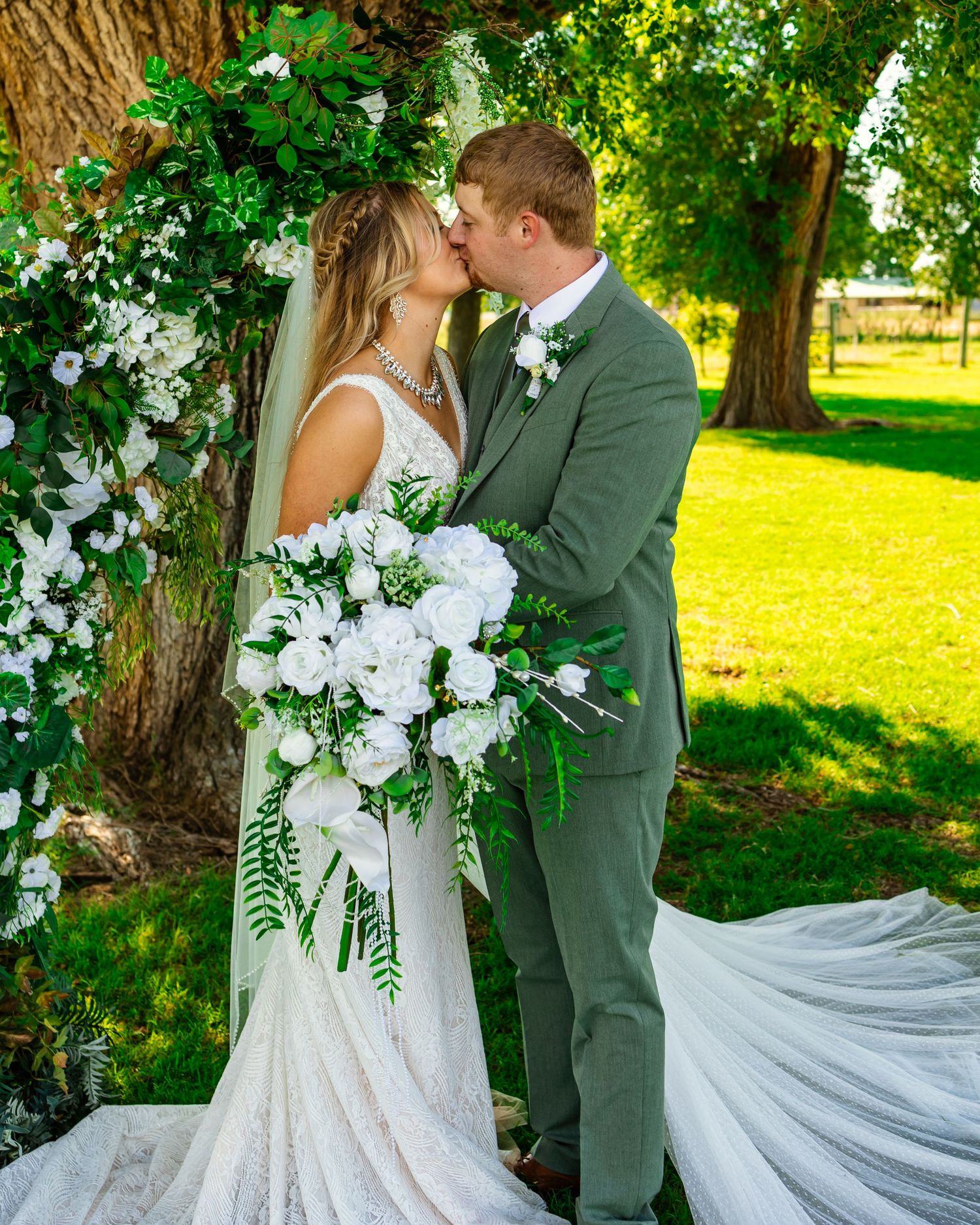 A bride and groom share a kiss under a floral arch with white roses and greenery on their wedding day at Darlington Chapel in El Reno, OK.