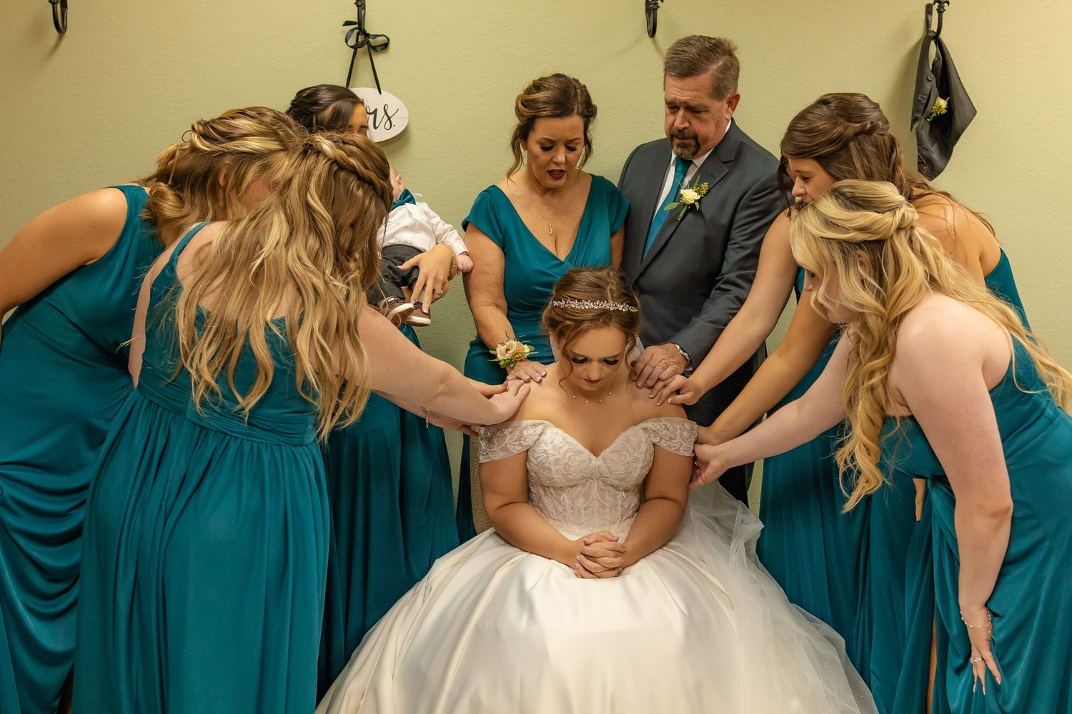 Bride in white gown surrounded by bridesmaids in teal dresses and groomsman praying together before wedding ceremony in oklahoma