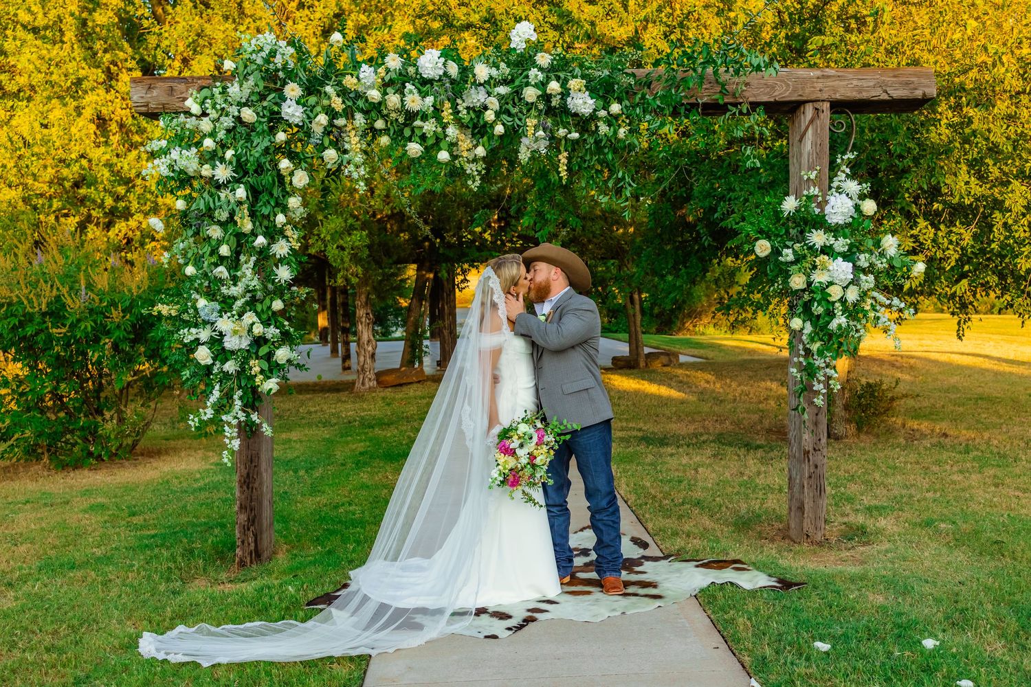 Bride and groom share first kiss under rustic floral arch with white roses at outdoor wedding ceremony at Rustic Rose Ranch in Guthrie, OK.