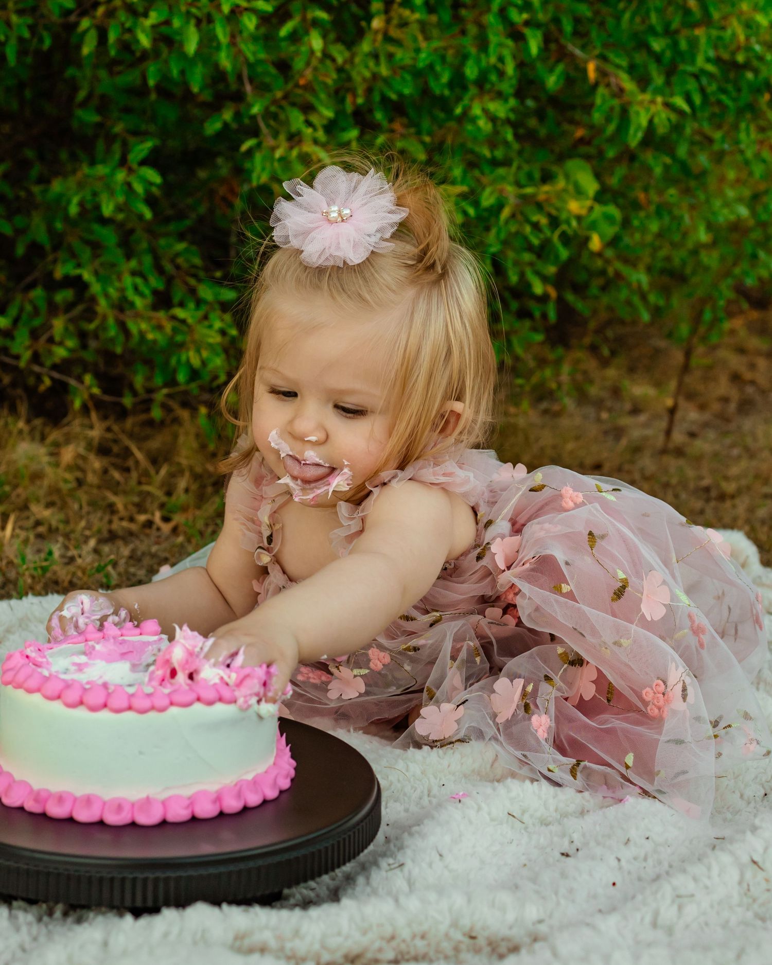 Toddler girl in pink tulle dress with flower hair clip smashing her first birthday cake outdoors in family photoshoot in oklahoma