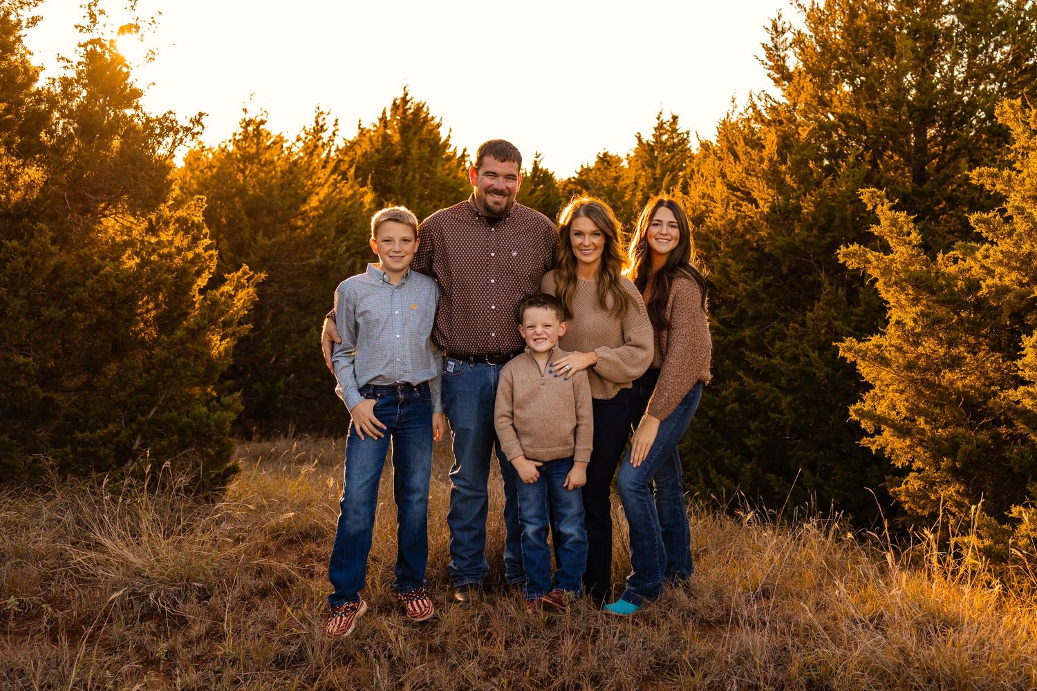 Family of five posing outdoors at golden hour in a field surrounded by trees, wearing coordinated casual outfits in Oklahoma family shoot