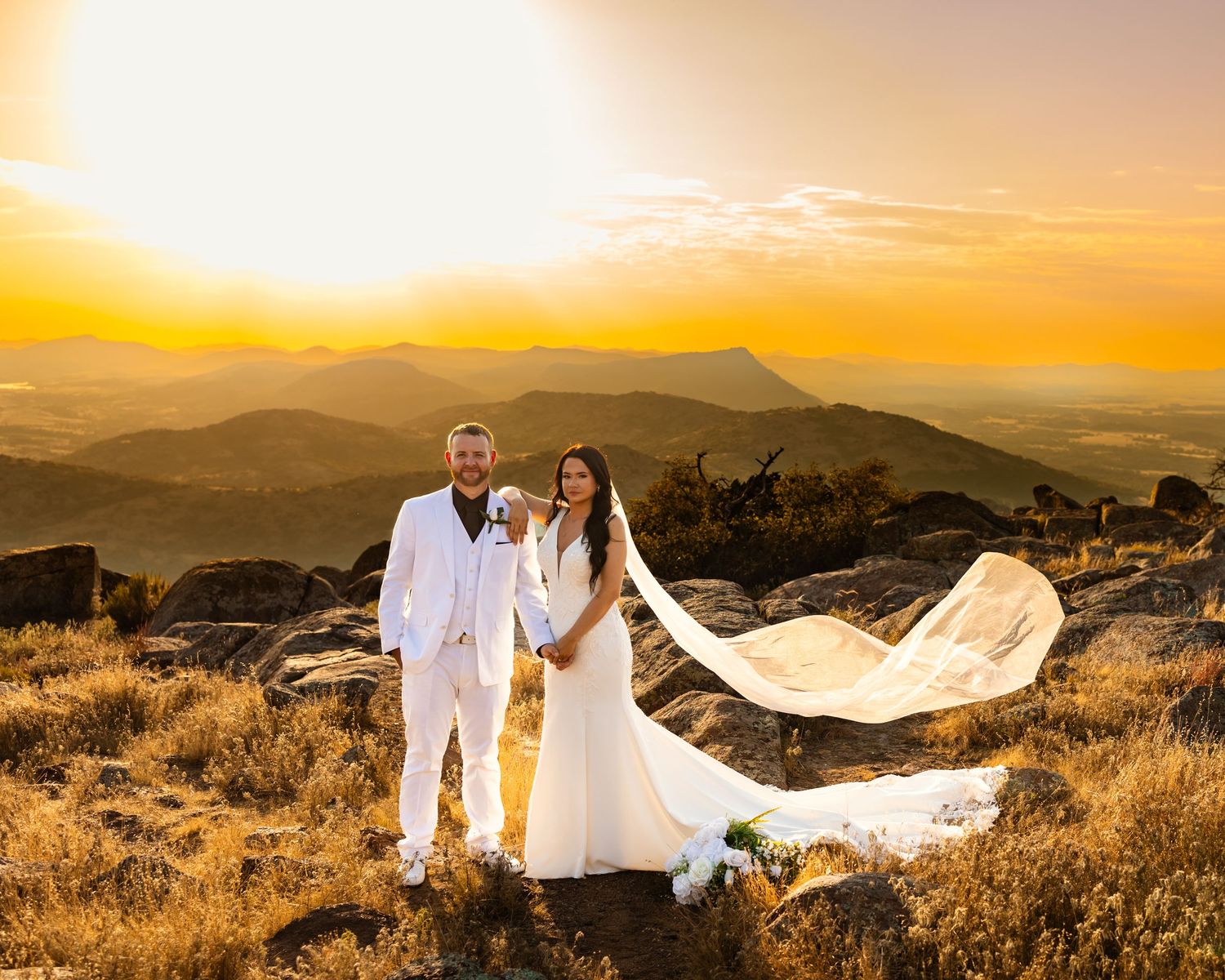 Bride and groom holding hands at golden sunset on an Oklahoma mountain top, veil flowing in wind, scenic valley backdrop.