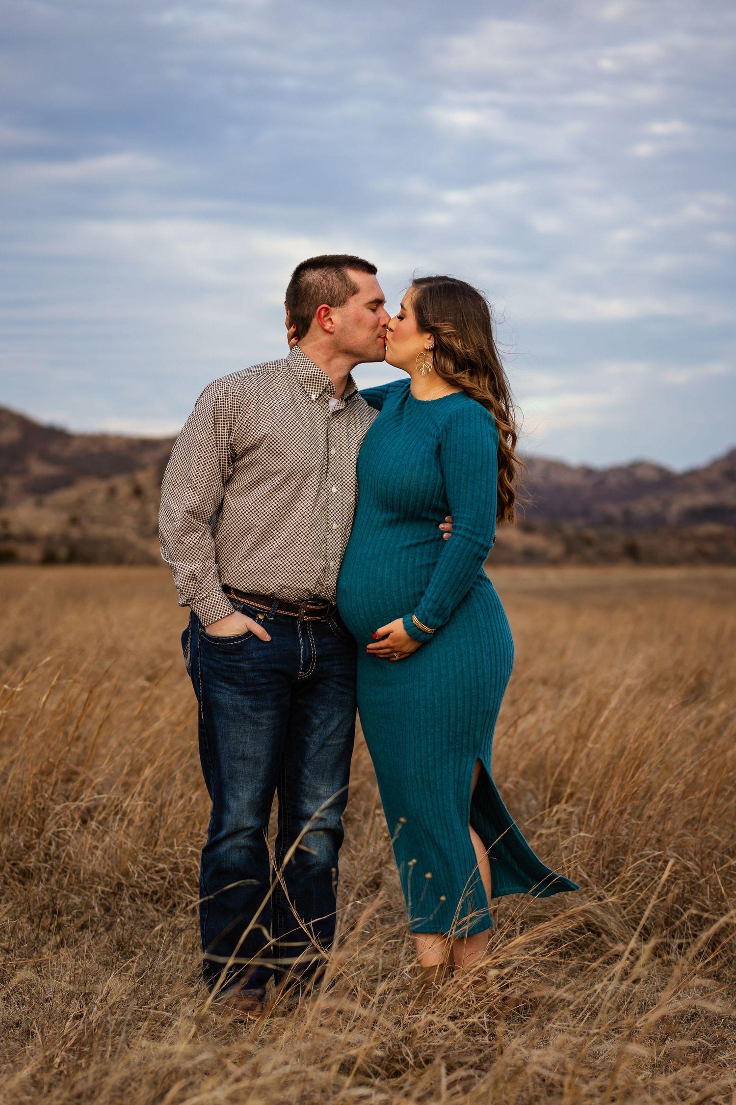 Couple sharing a kiss in a golden field, woman in teal maternity dress cradling baby bump, mountains in background. Oklahoma maternity