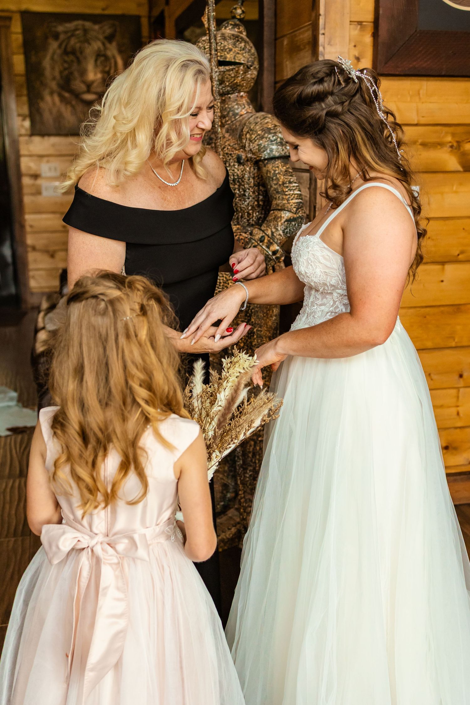 Bride in white gown shares a tender moment with flower girl and woman in black dress at rustic wooden venue in oklahoma