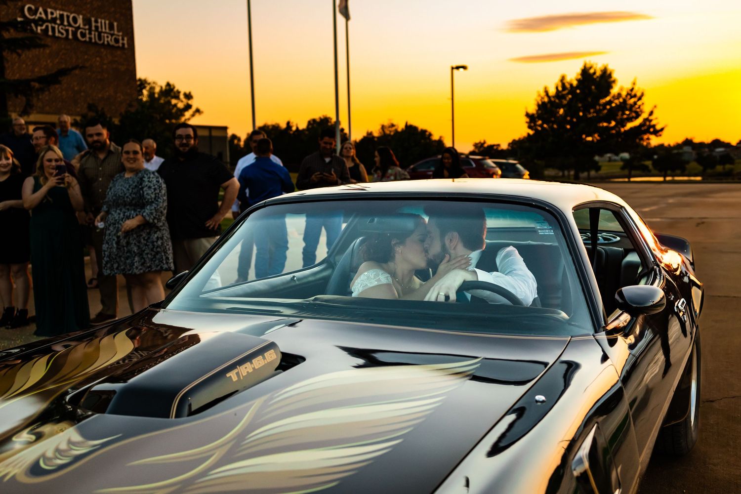 Couple sharing a kiss in a classic black muscle car at sunset outside a church with wedding guests watching in oklahoma