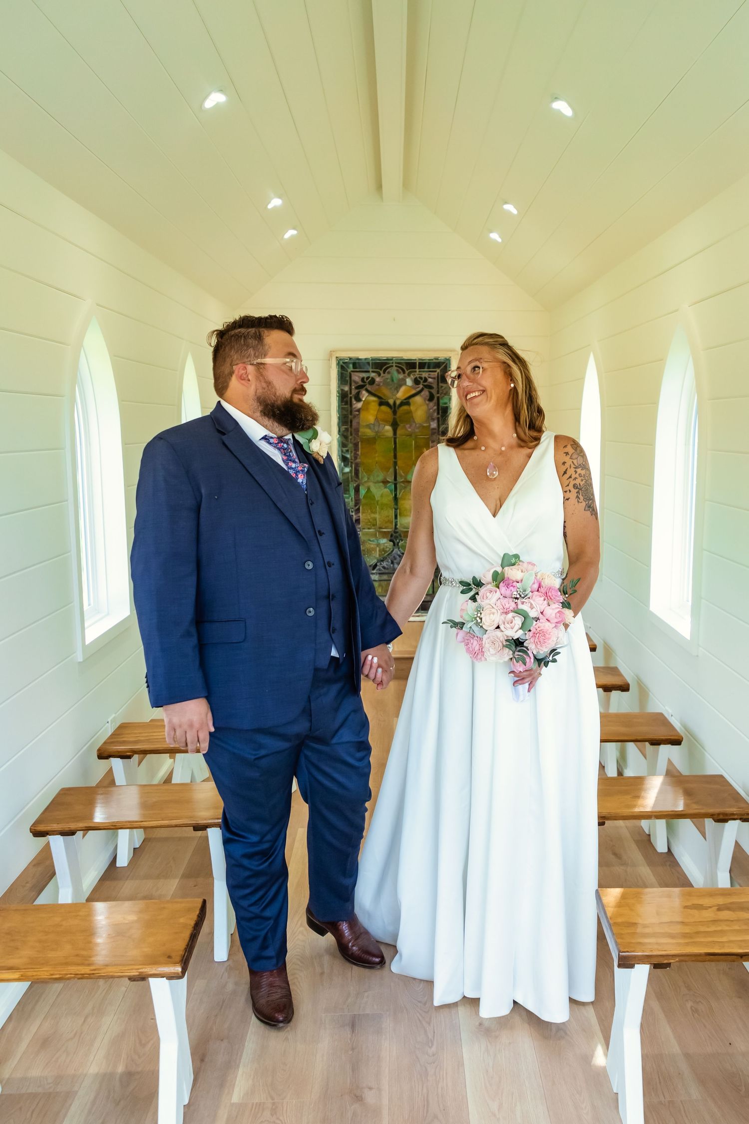 Bride and groom holding hands and smiling in a small white chapel aisle, bride holding pink bouquet at hitched traveling wedding chapel
