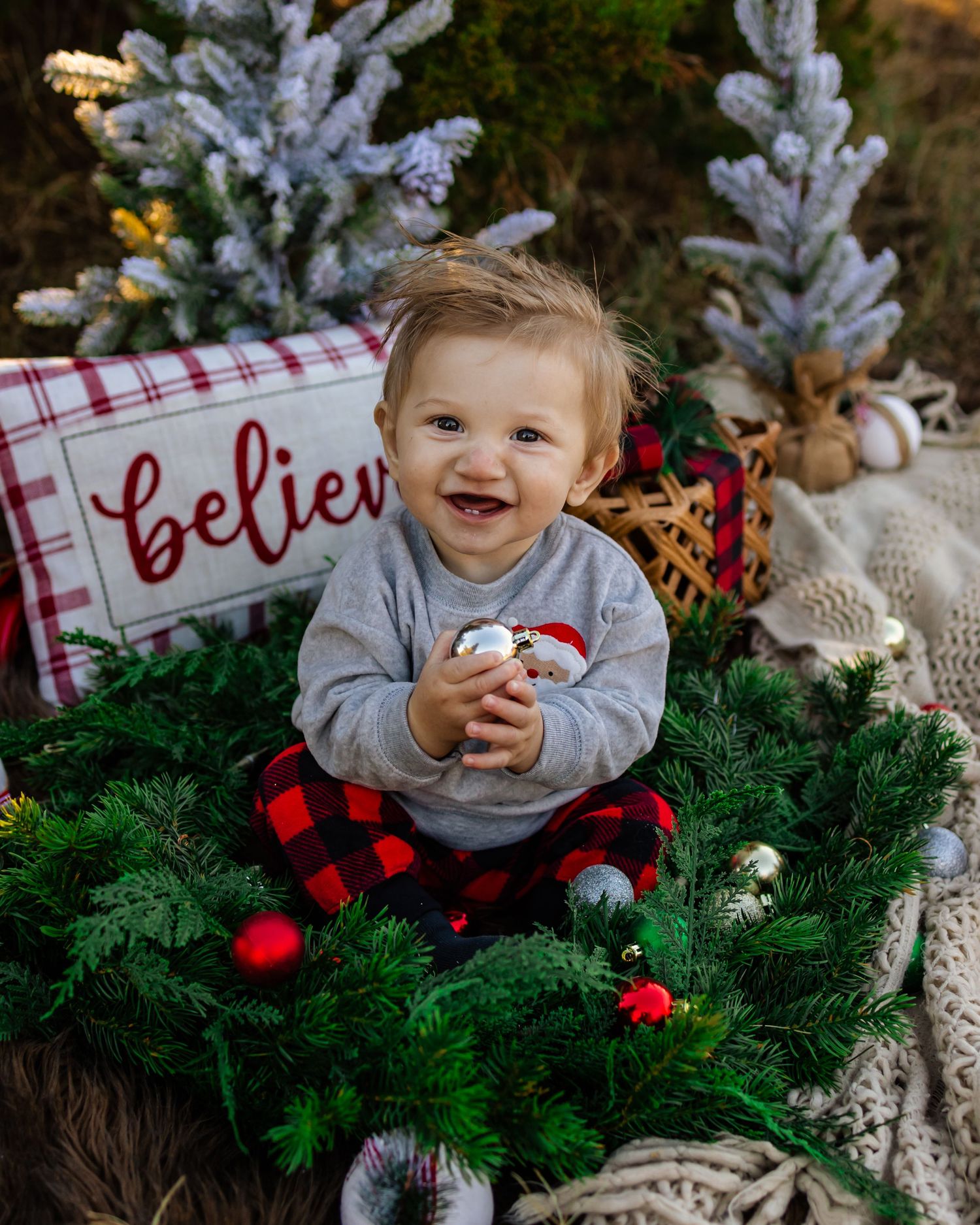 Smiling toddler in red plaid pants sits on Christmas wreath surrounded by festive decorations and a Believe sign in Oklahoma photography