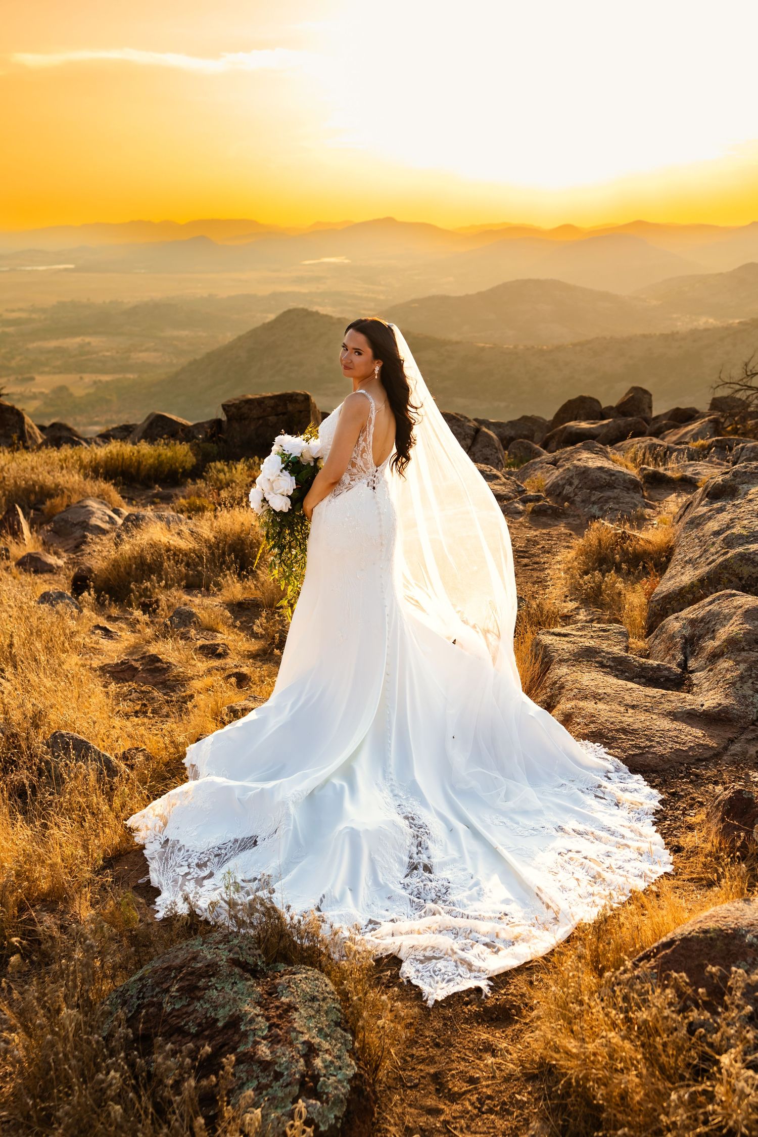 Bride in white gown and veil stands on rocky mountain at golden sunset, holding white bouquet with scenic valley views in Oklahoma elopement