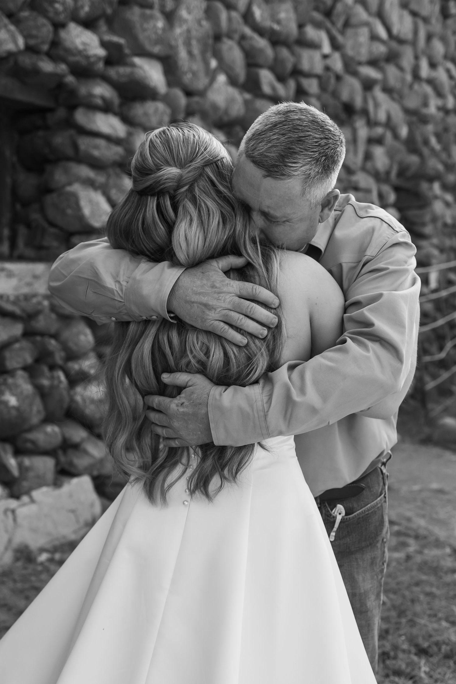 Black and white photo of a dad embracing his daughter in a wedding dress near a stone wall. Oklahoma first look with dad