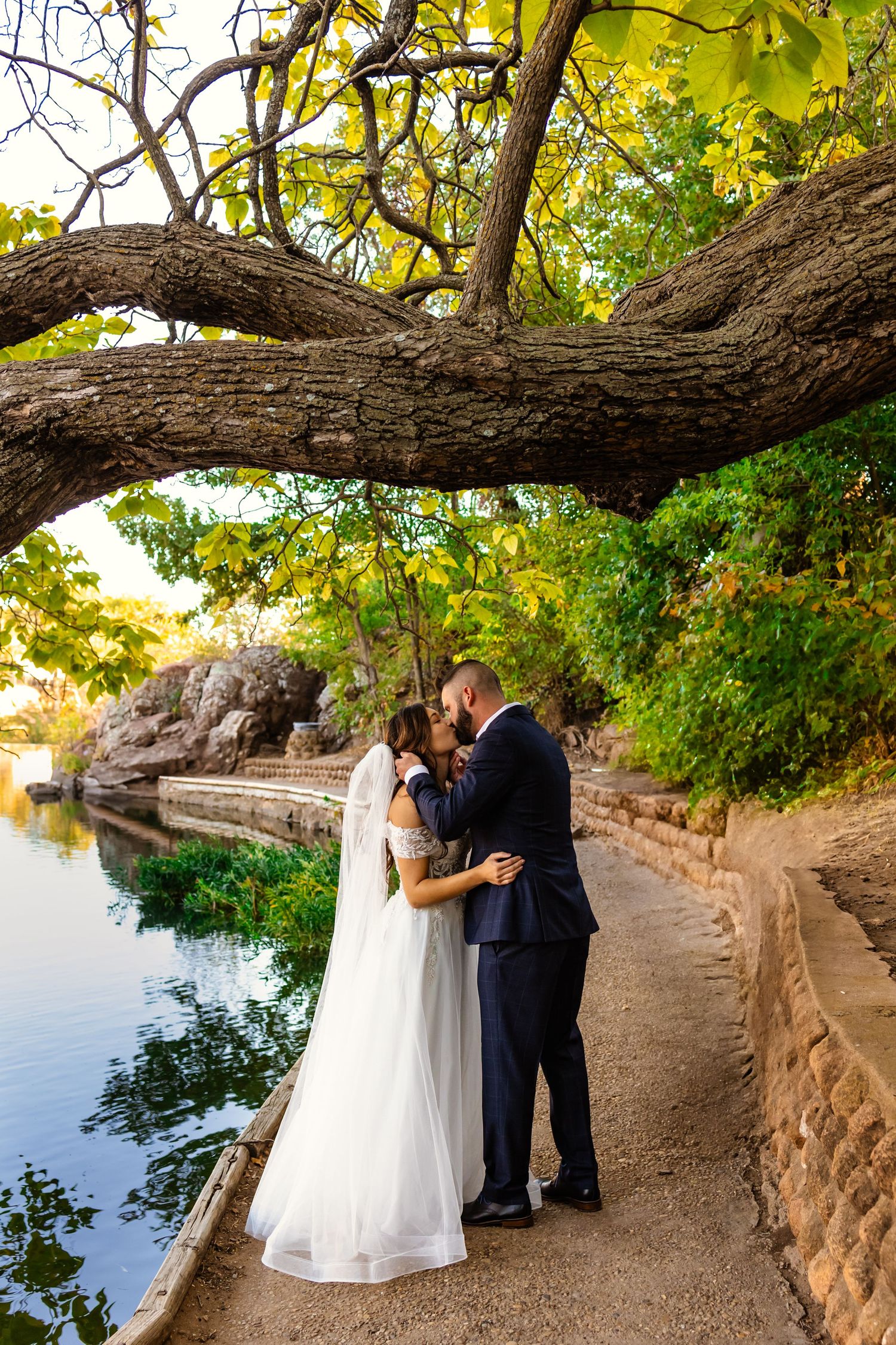 Bride and groom kissing under a large tree branch beside a serene river in a lush outdoor wedding elopement in medicine park, oklahoma