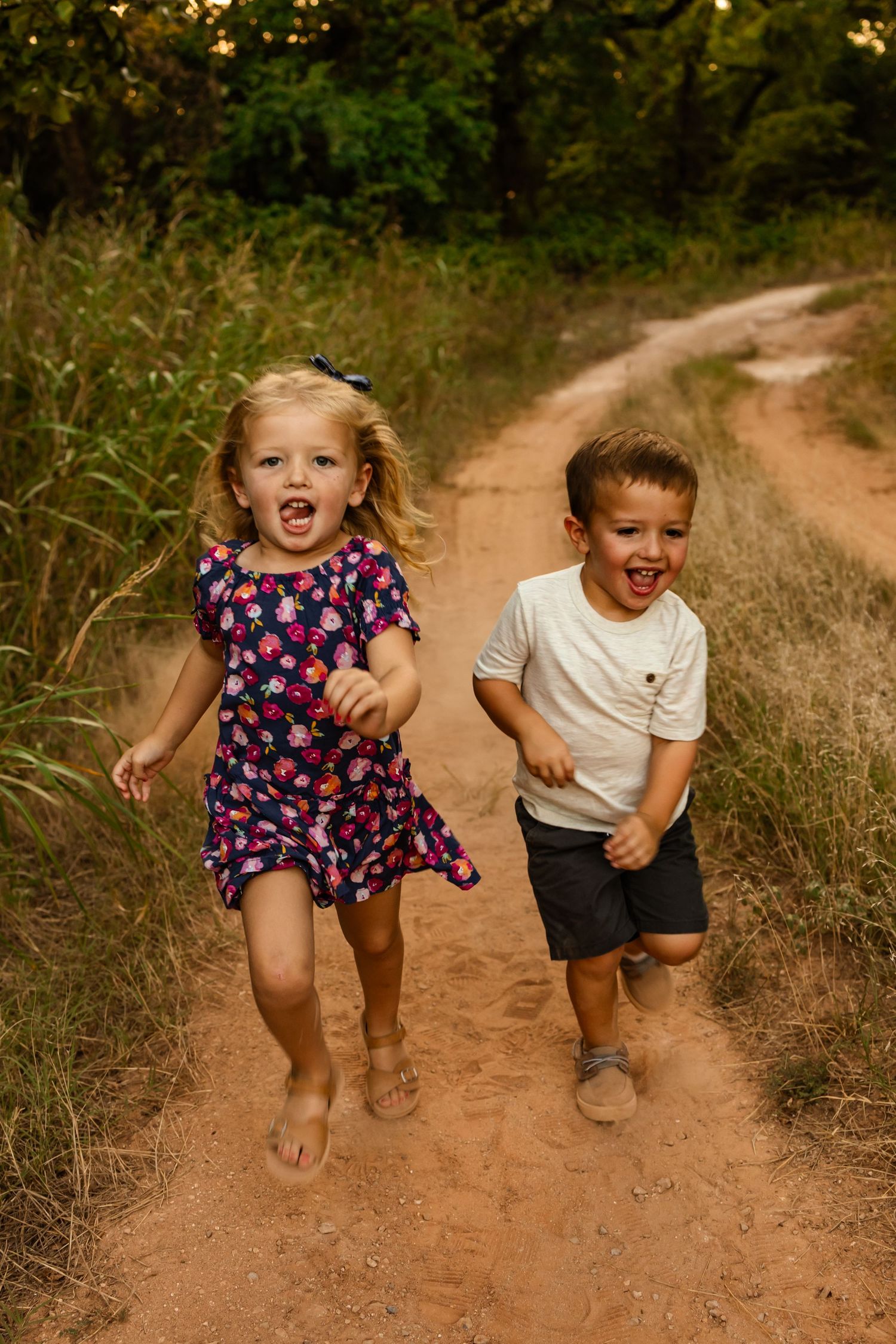 Two young children laughing and running together on an oklahoma dirt path surrounded by lush green trees. family session
