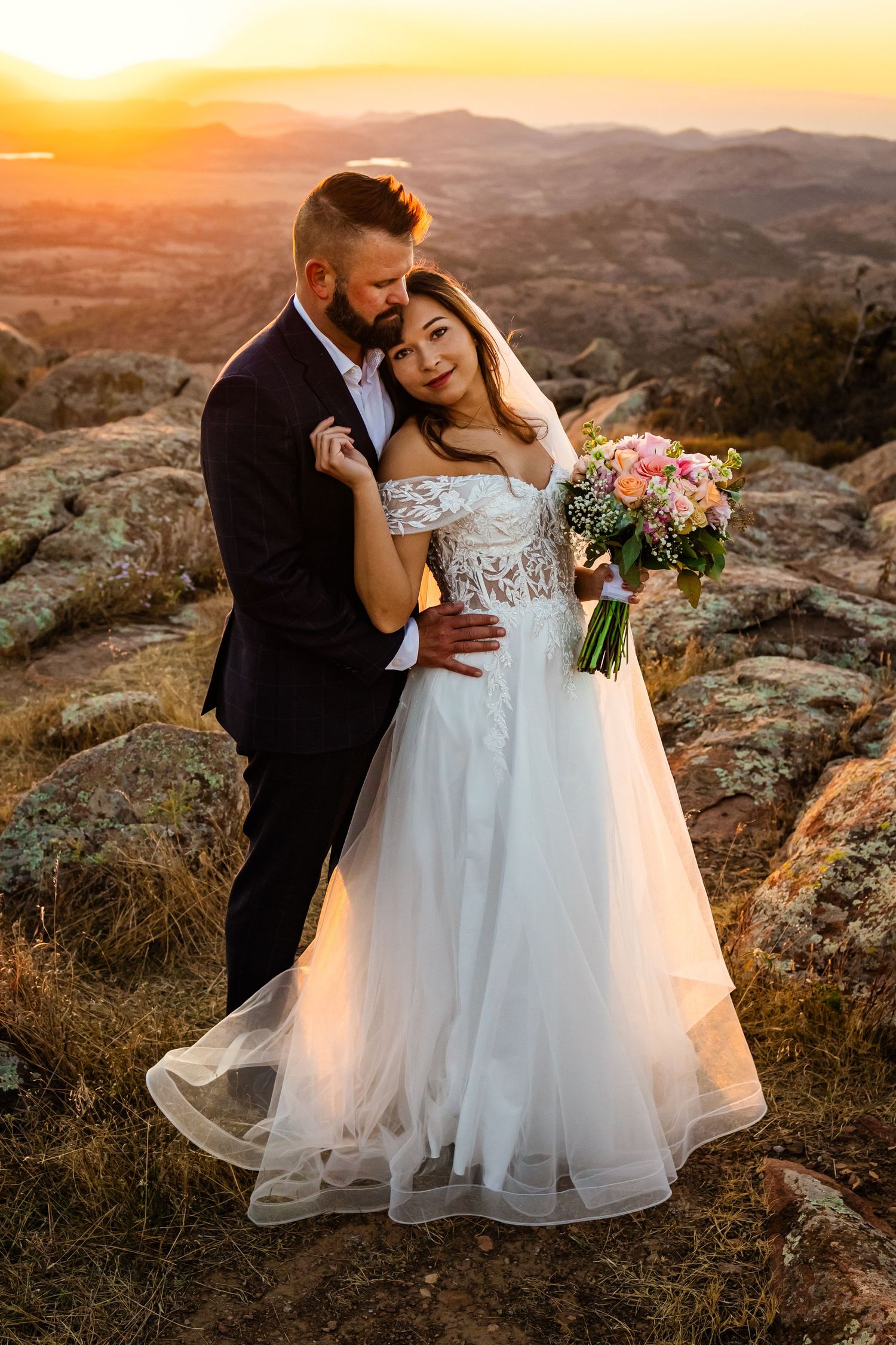 Bride and groom embrace at golden sunset on rocky mountaintop, bride holding colorful bouquet in white tulle gown mount Scott oklahoma