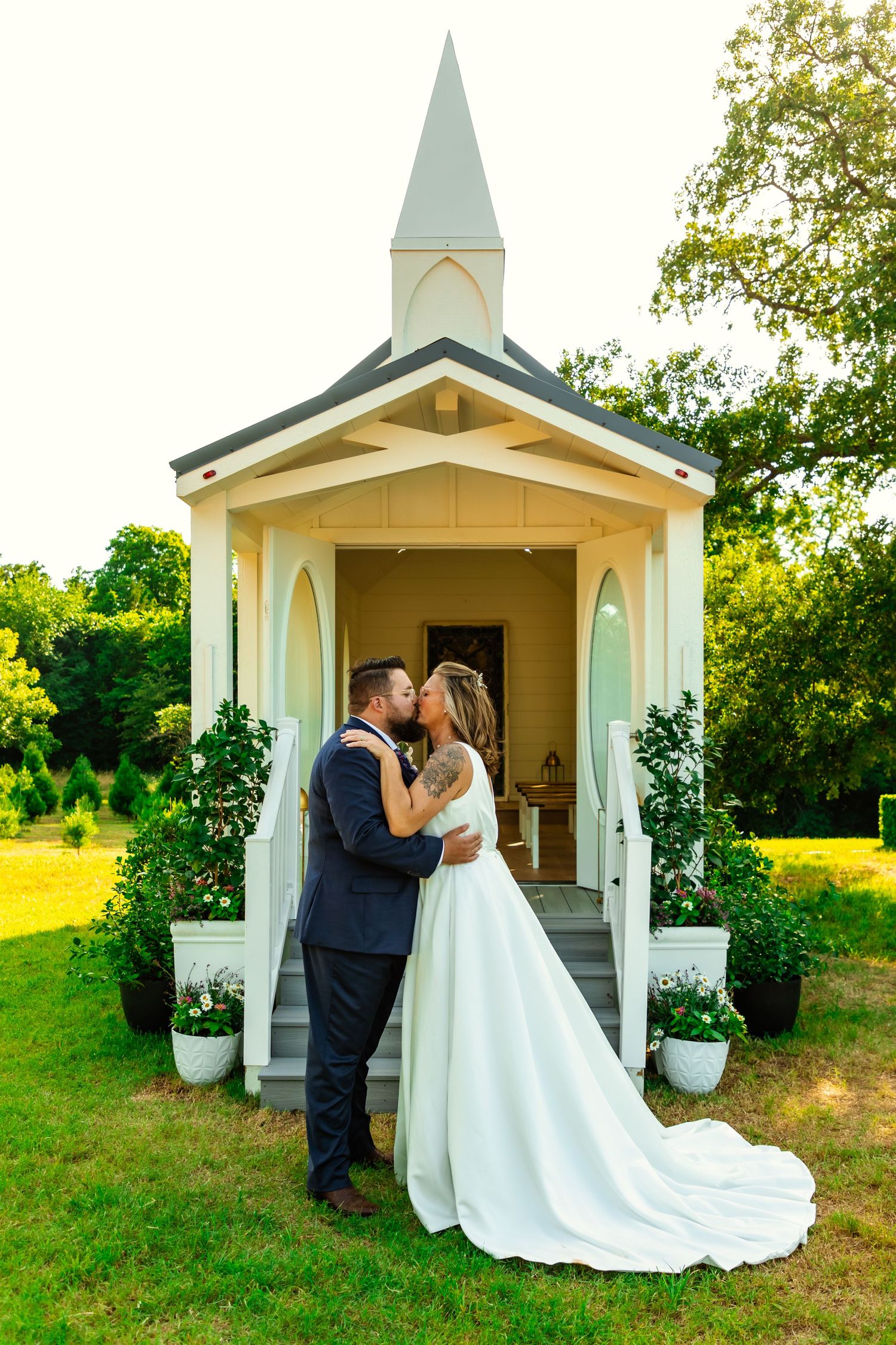 Bride and groom kissing in front of a charming mini chapel surrounded by greenery on their wedding day. hitched traveling wedding chapel, ok