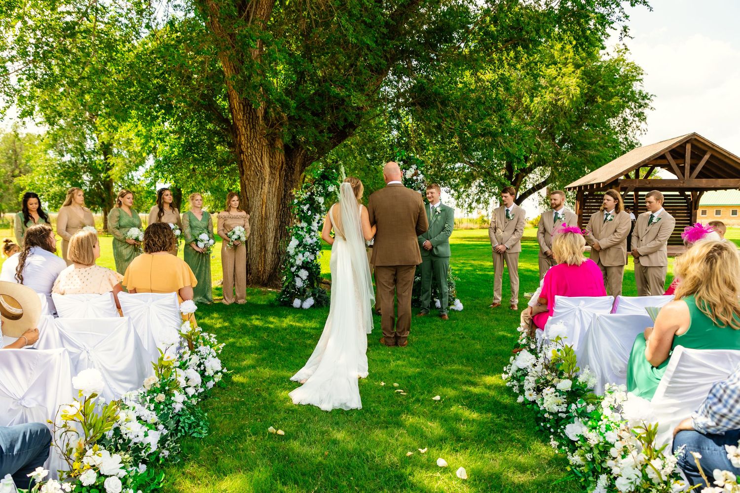 Outdoor wedding ceremony on lush green lawn under large tree with floral aisle and guests seated on white chairs. darlinton chapel, OK