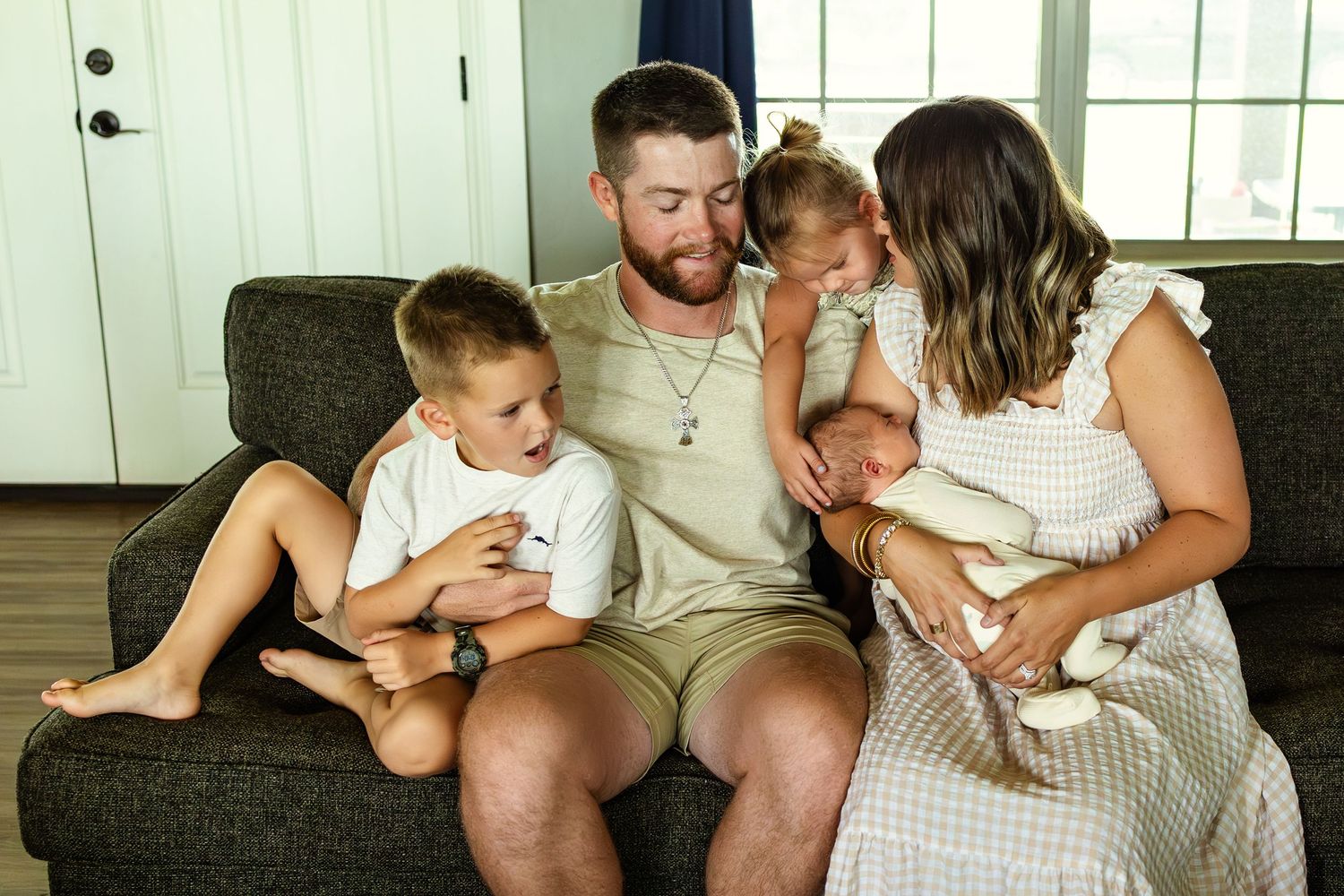 Happy family of four sitting on a gray couch, parents smiling at newborn baby with young boy beside them in family session in oklahoma