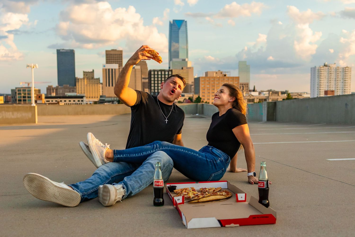 A couple sits on a rooftop parking lot enjoying pizza and Coca-Cola with a Oklahoma city skyline at sunset behind them.