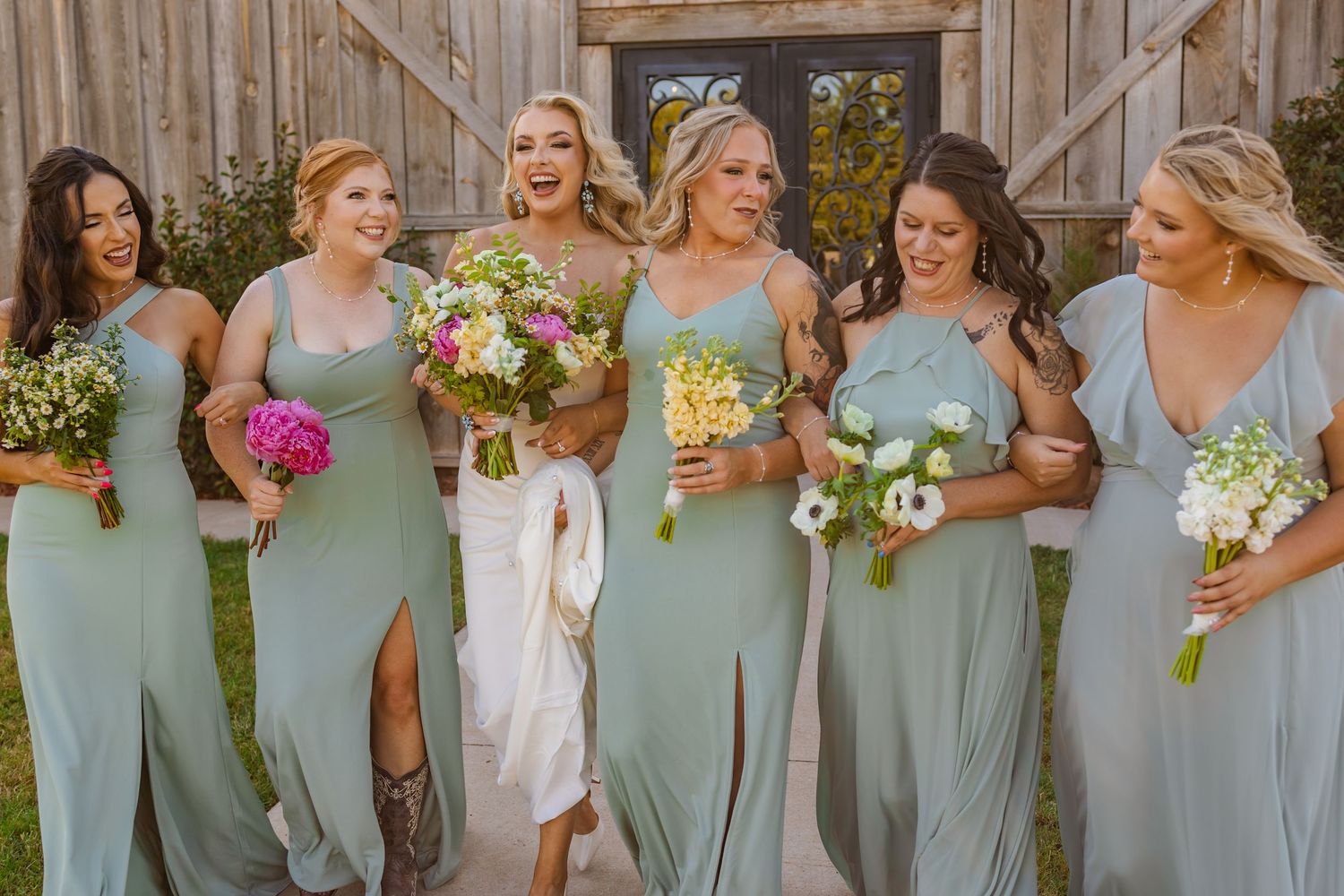 Six bridesmaids in sage green dresses laughing and walking together outside at rustic rose ranch holding colorful bouquets.