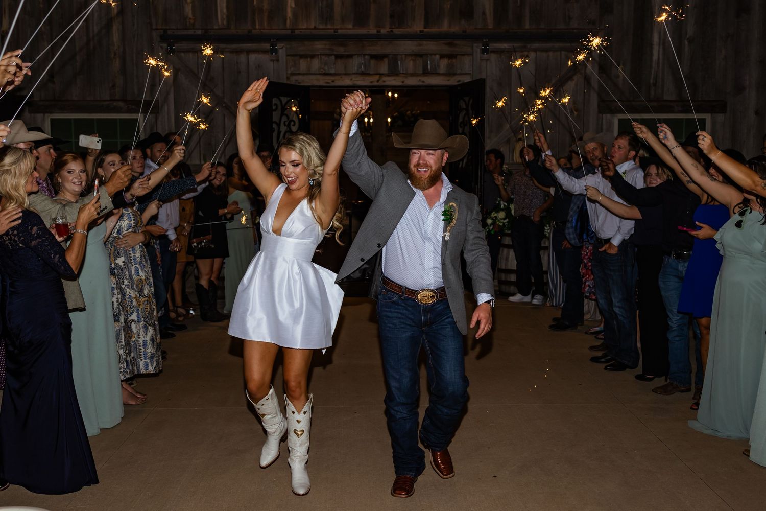 Bride in white mini dress and boots exits barn wedding with groom in cowboy hat amid sparklers at rustic rose barn in Guthrie Oklahoma.