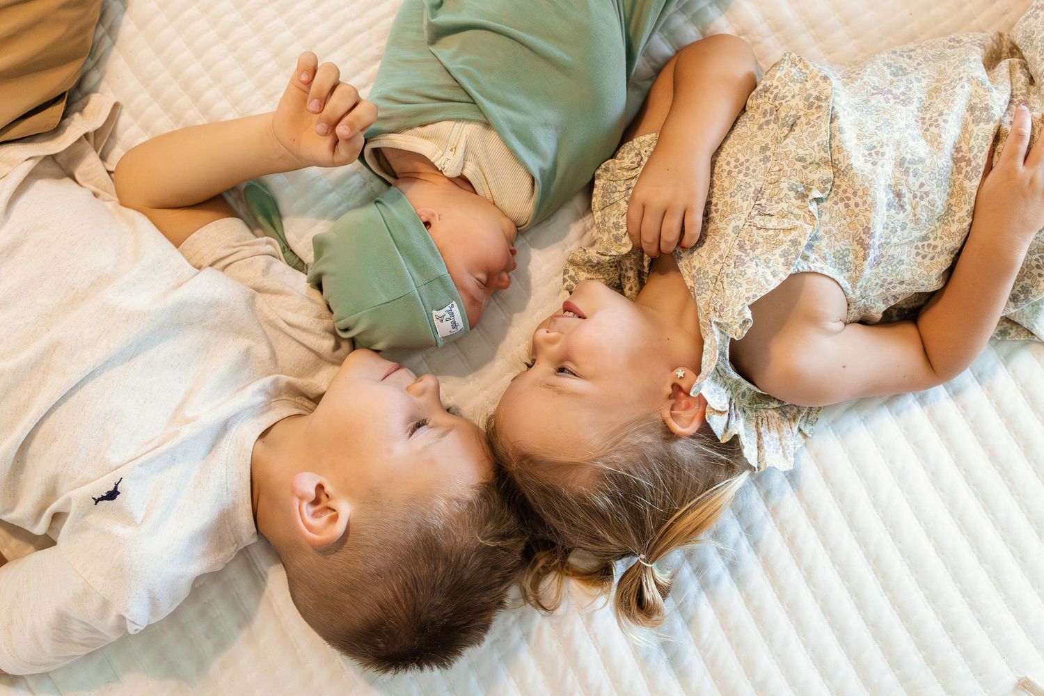 Three young children lying together on a white bed, smiling and bonding in a sweet, candid moment in home Oklahoma family session