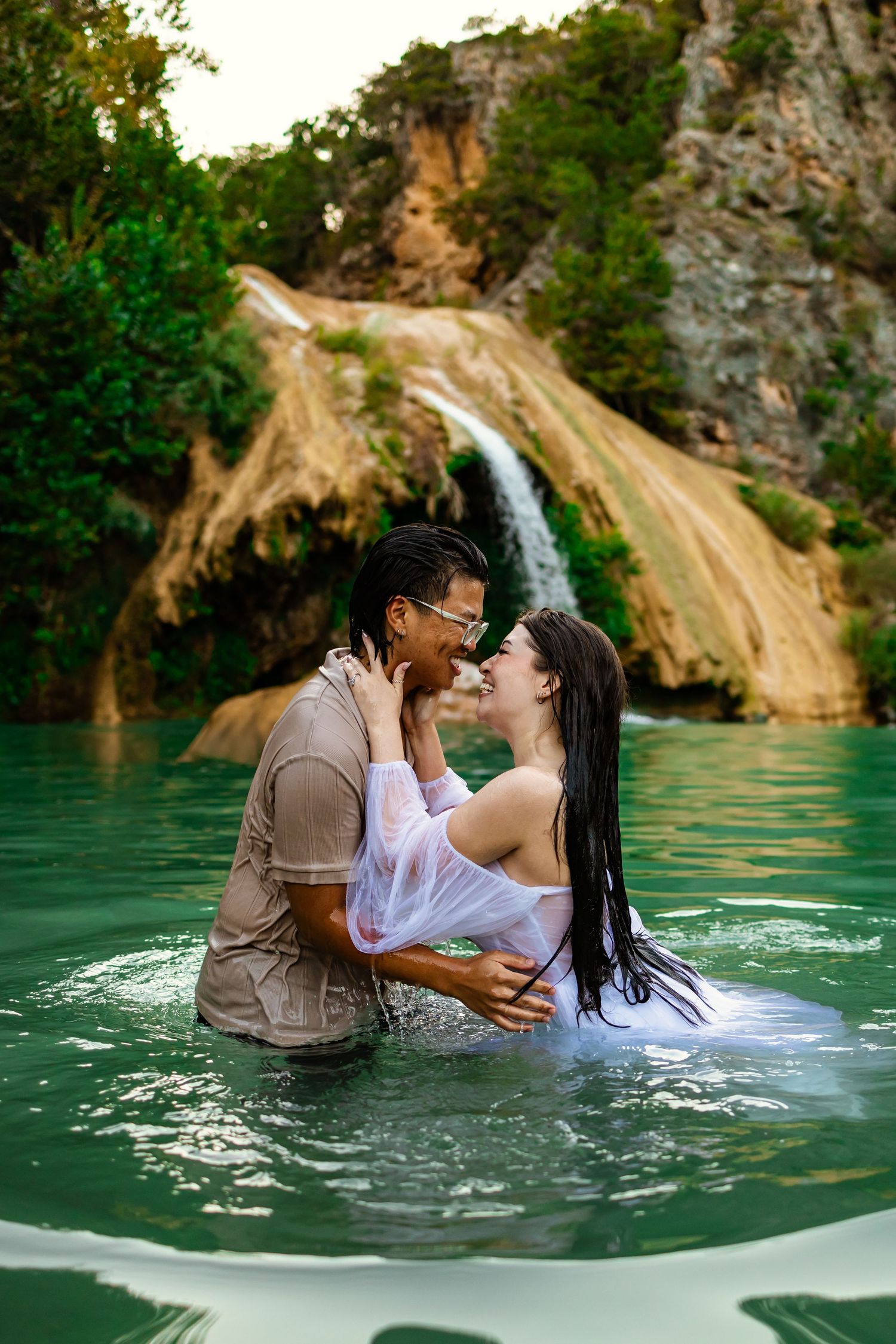 Couple sharing a romantic moment in a turquoise pool with a scenic waterfall and lush greenery in the background at Turner falls in Oklahoma