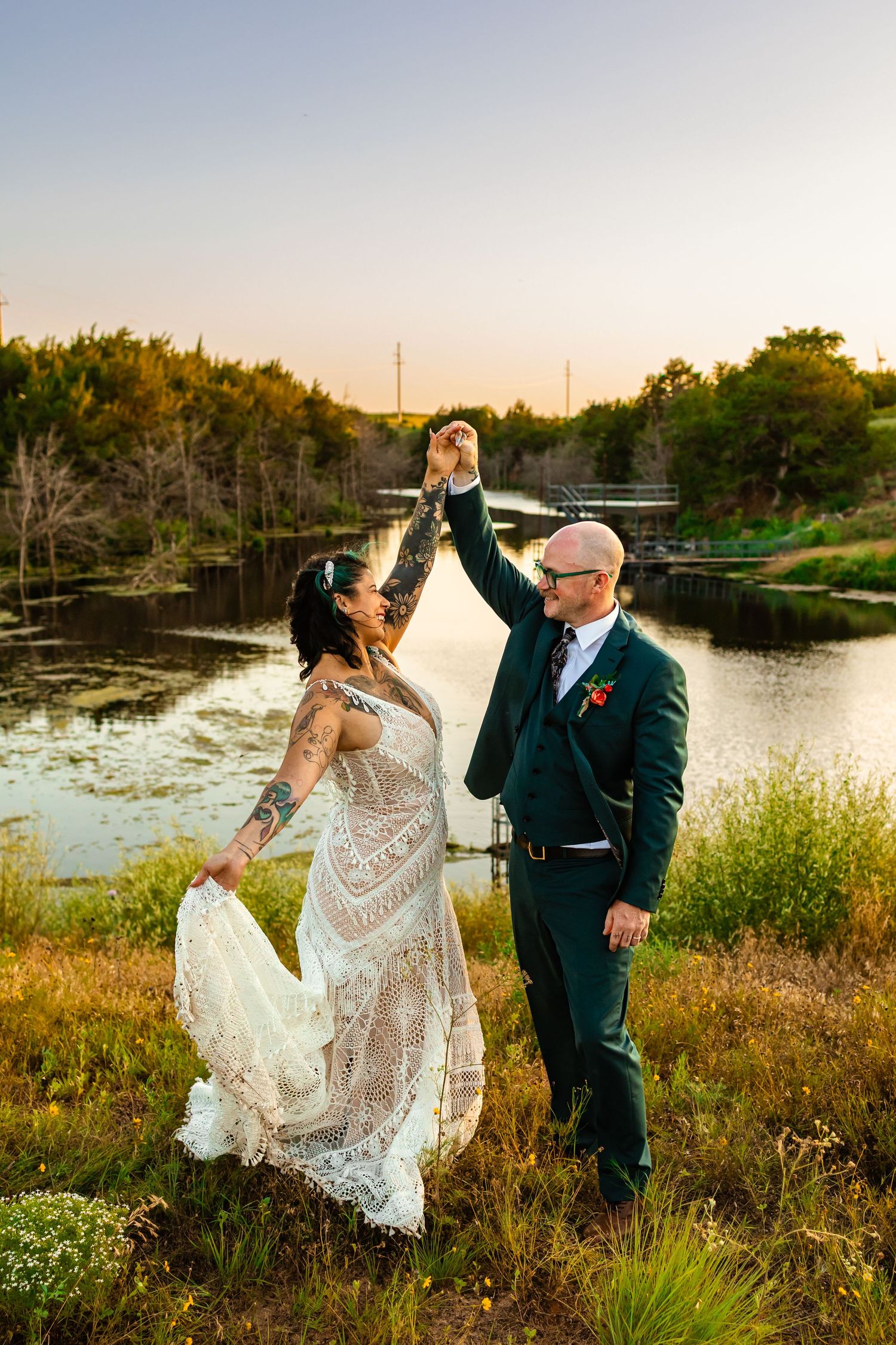 Bride and groom dancing outdoors at sunset near a serene river, bride in lace gown, groom in green suit at Weatherford, Oklahoma wedding.