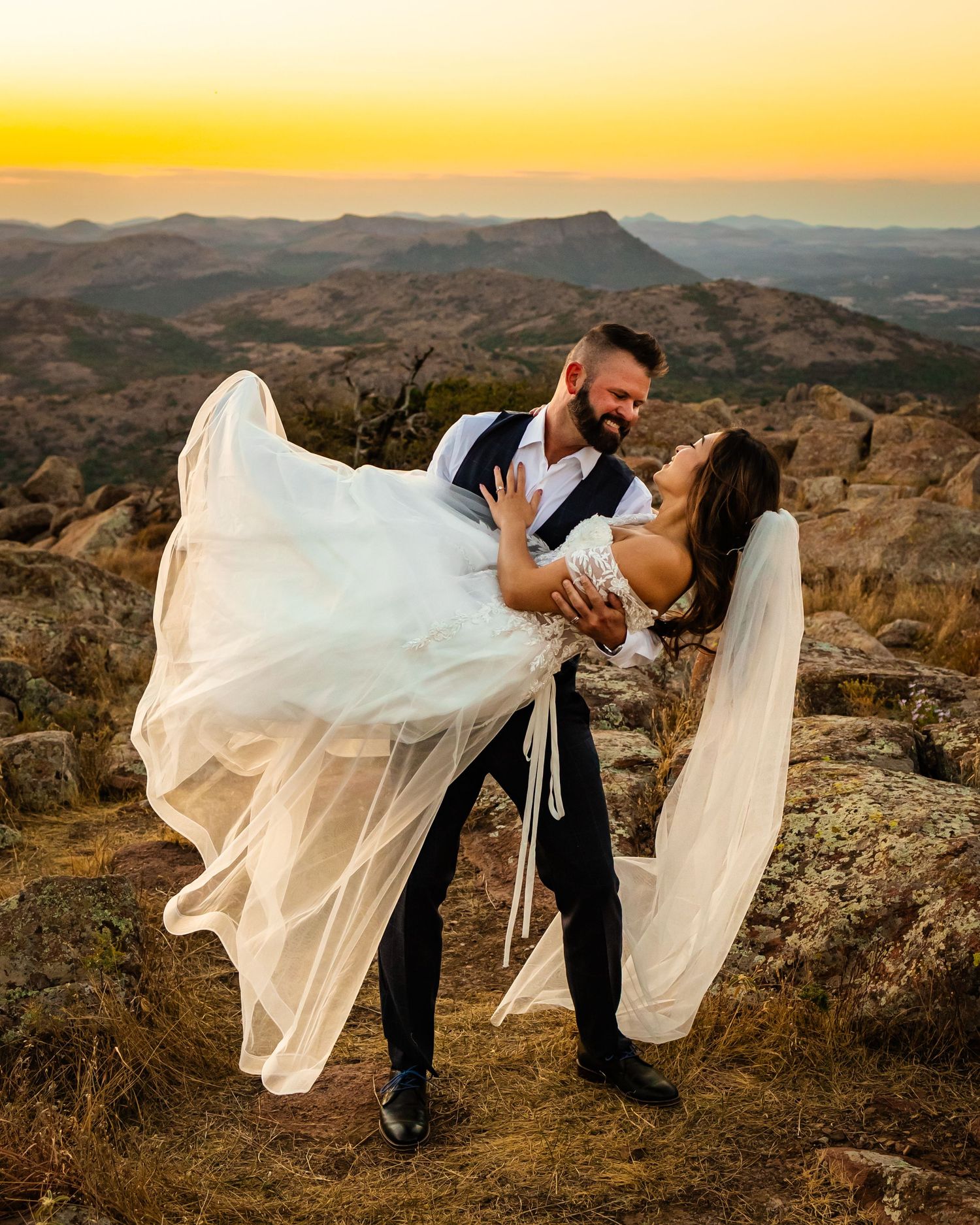Groom lifting bride in white gown at sunset on rocky mountain overlook with Oklahoma golden sky backdrop.