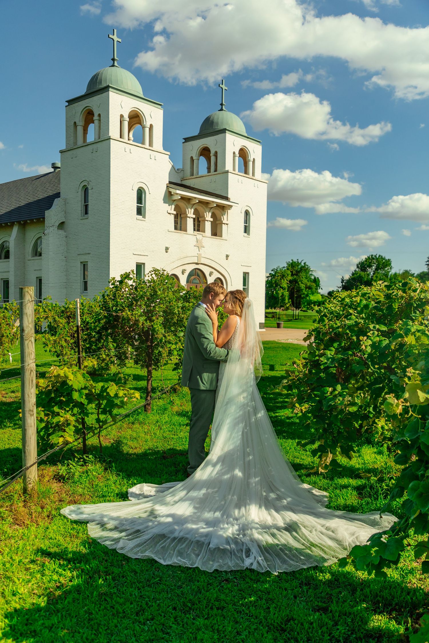 Bride & groom embrace in vineyard before white church with twin towers and crosses on sunny wedding day at darlington chapel in el Reno, OK