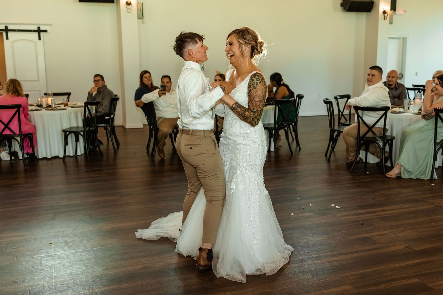 Bride and bride share their first dance at wedding reception, bride in white gown, bride in tan suit, guests seated nearby at sorelle in OK