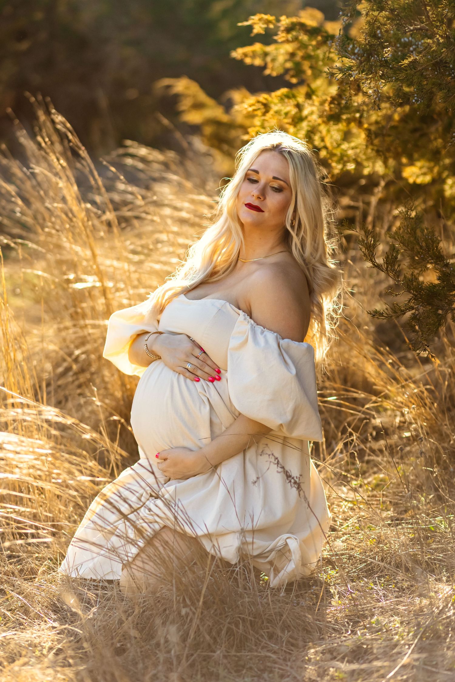 Pregnant woman in cream off-shoulder dress kneeling in golden Oklahoma field, cradling baby bump in warm sunset light.