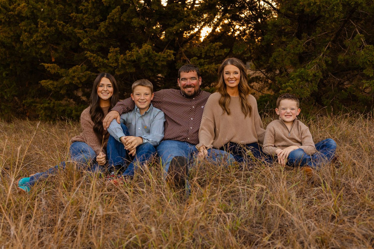 Family of five smiling together in golden field at sunset in oklahoma, surrounded by trees in warm autumn light.