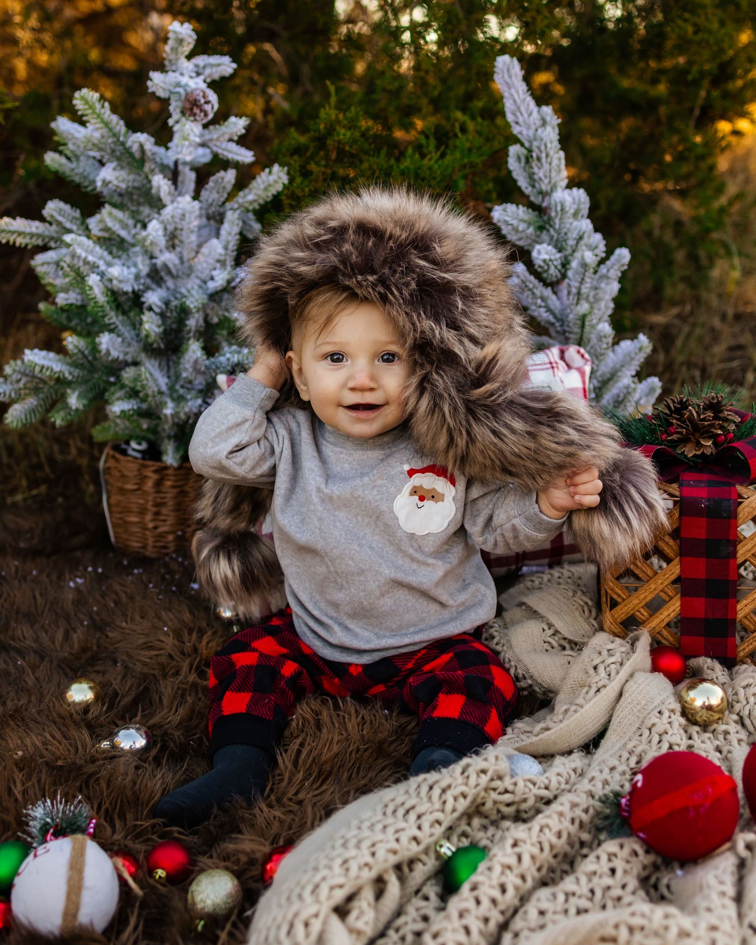 Adorable baby in Santa shirt, fur hood, and plaid pants sits among Christmas trees and festive decorations outdoors in oklahoma