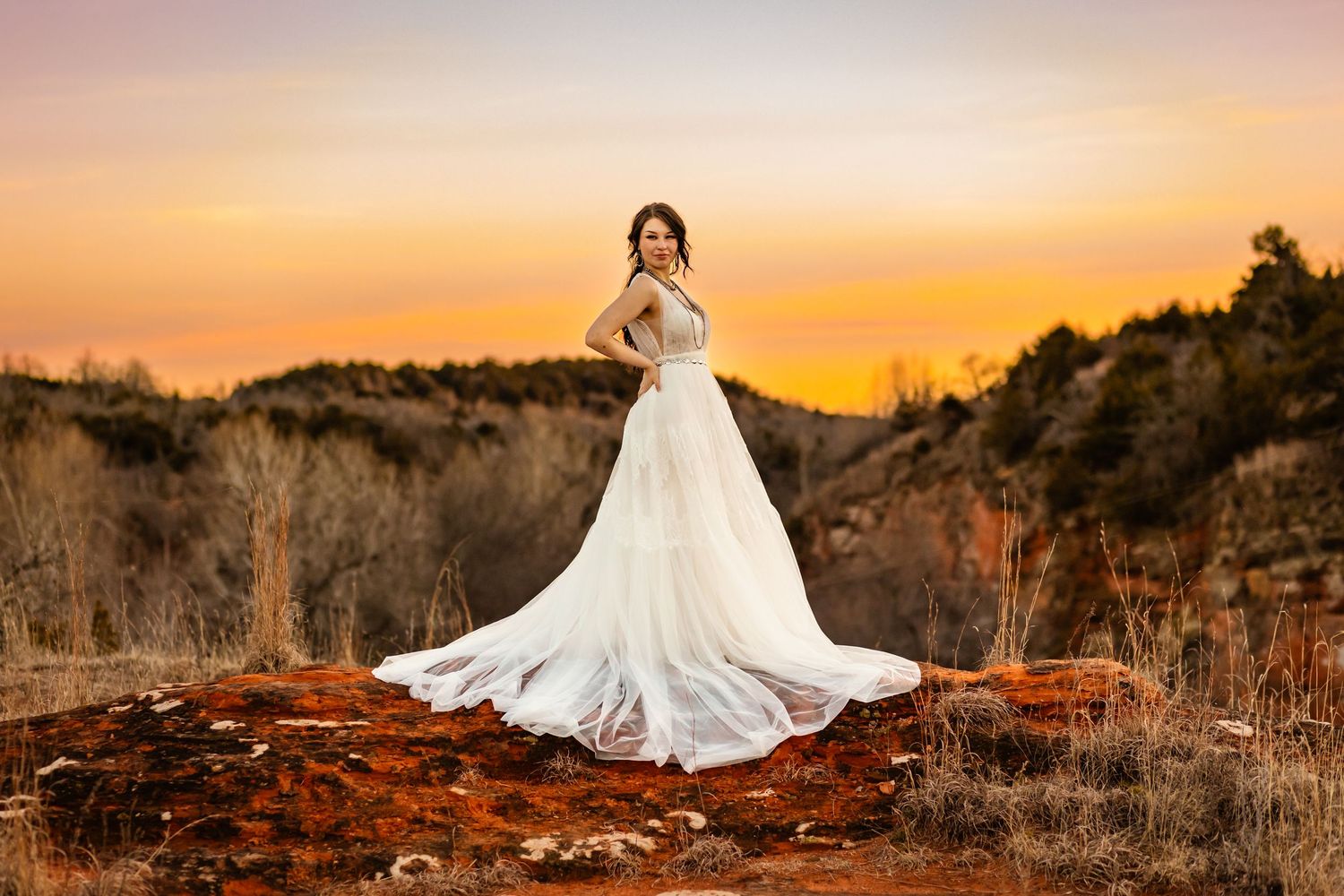 Bride in white gown with long train stands on red rocks at red rock canyon in Hinton Oklahoma at sunset in a scenic outdoor landscape.