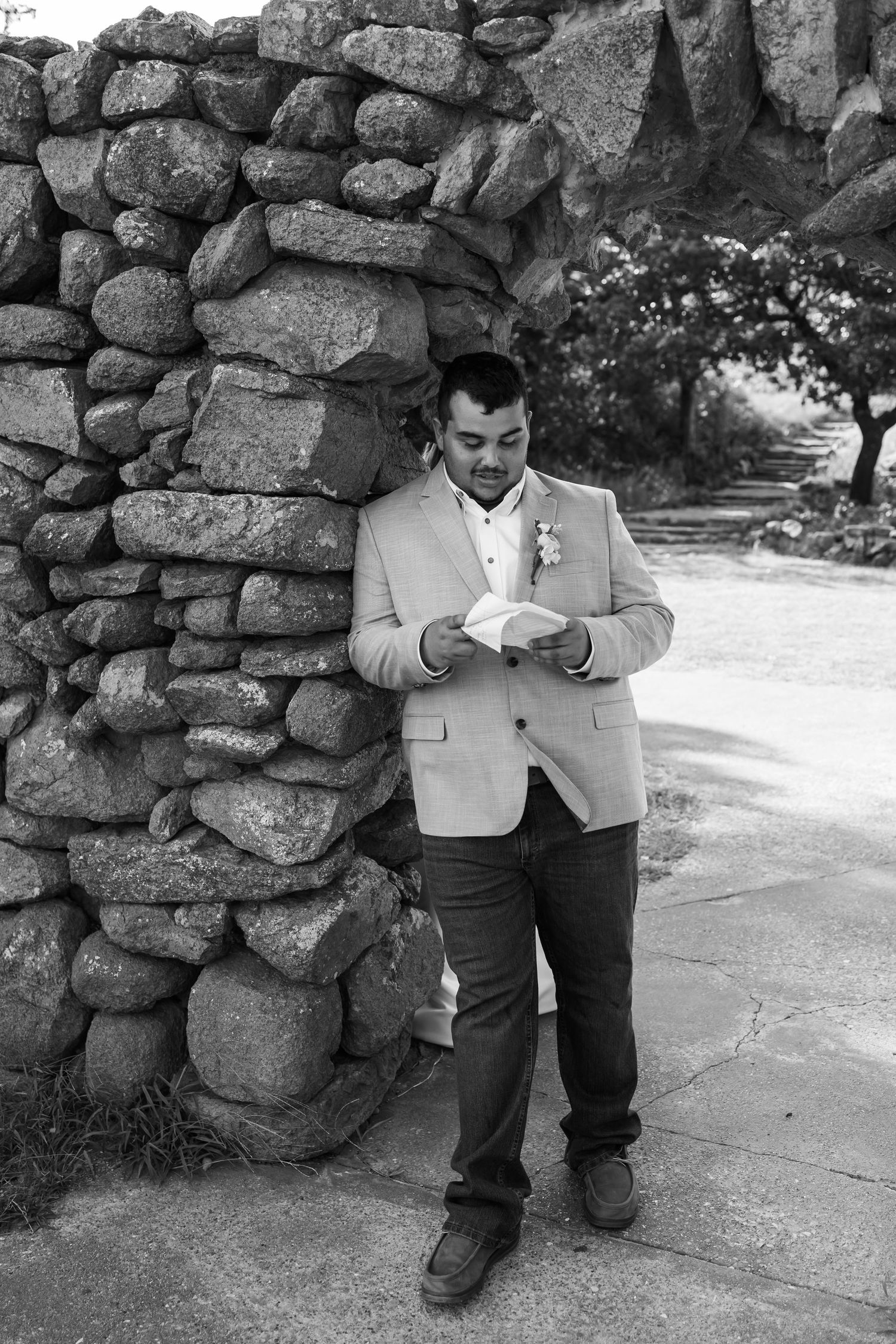 Groom in light blazer reads vows beside rustic stone wall, black and white oklahoma wedding portrait outdoors.