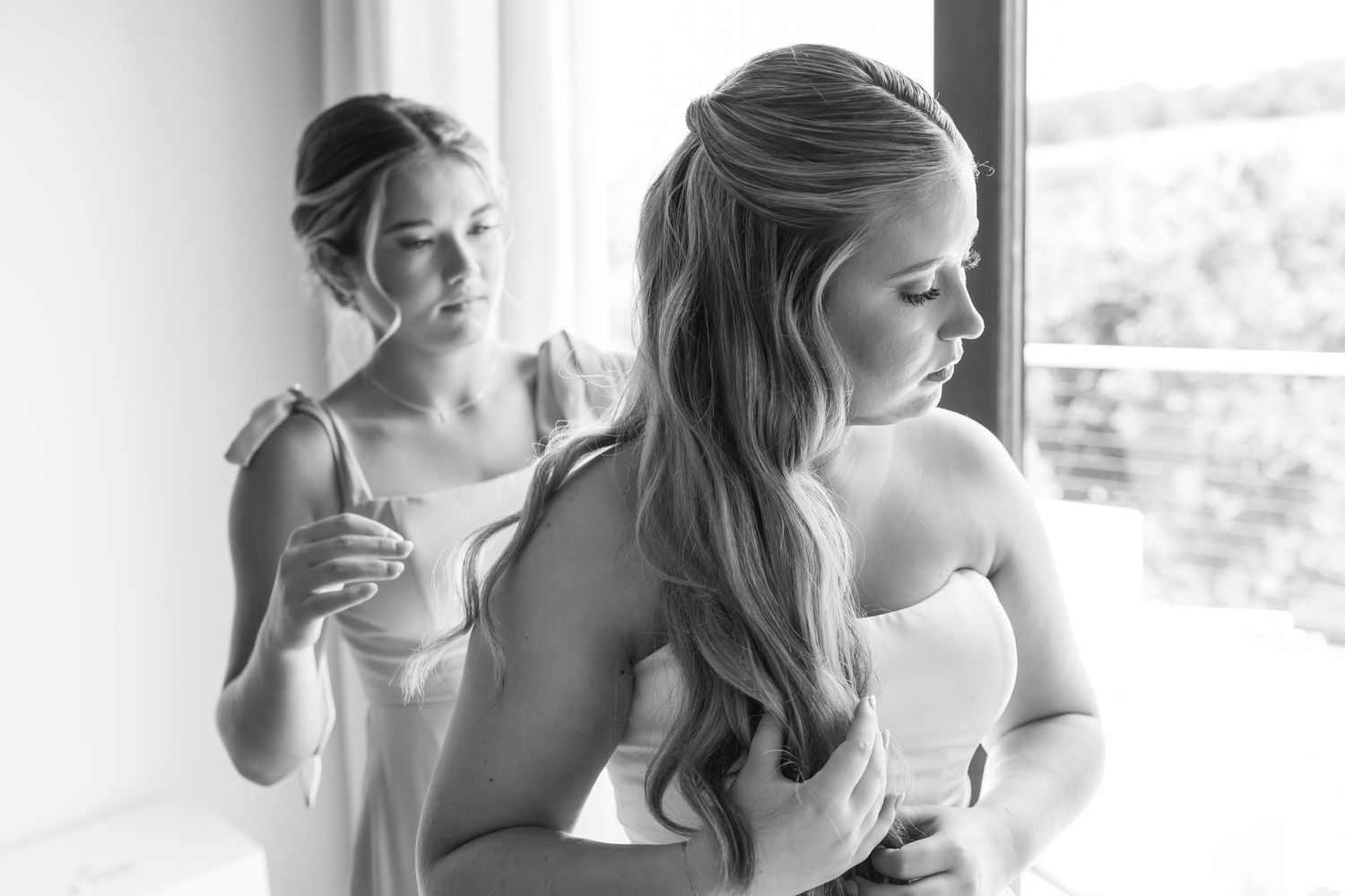 Bridesmaid helping Oklahoma bride with wedding dress in elegant black and white photo near bright window.