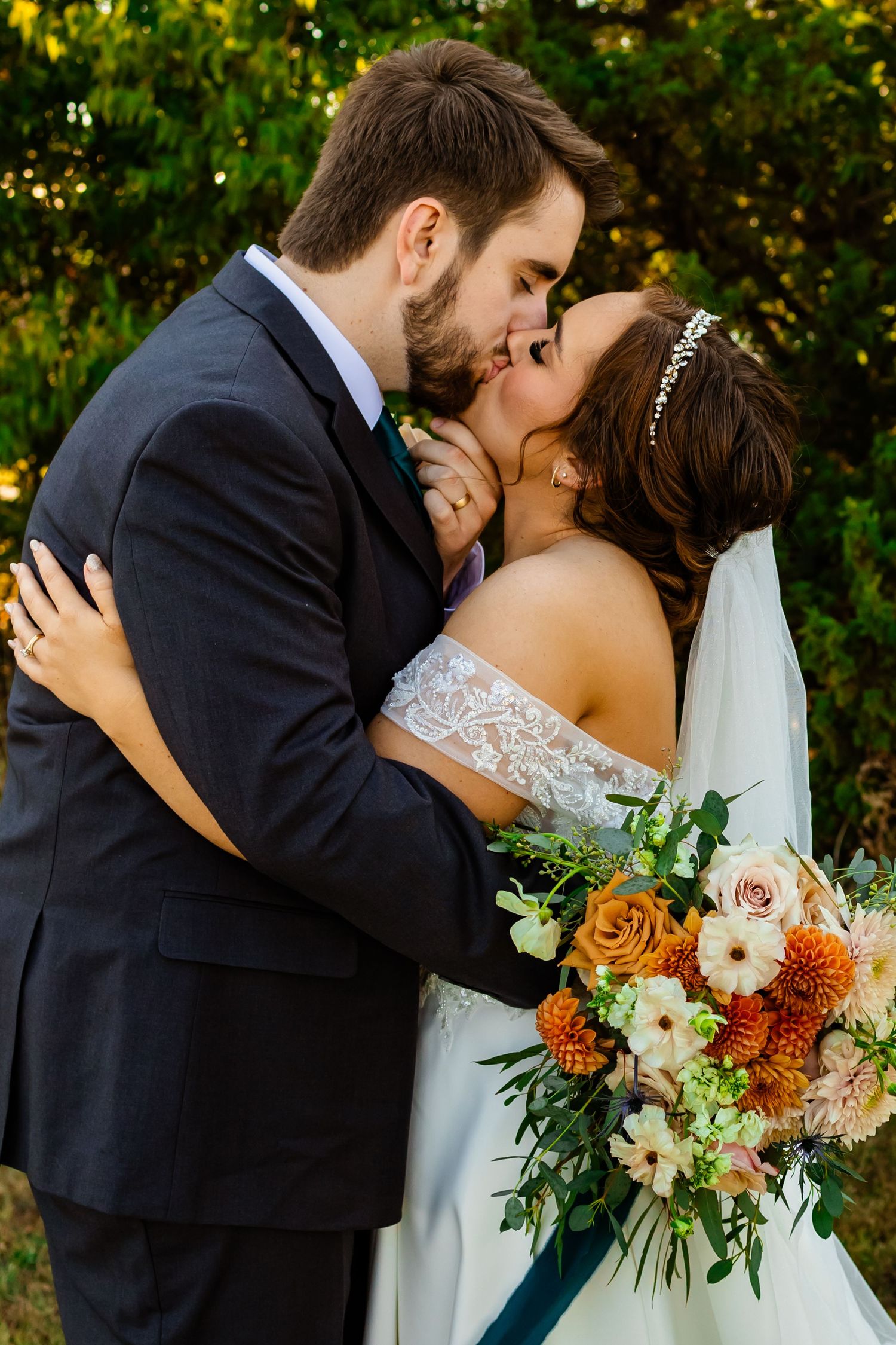 An oklahoma bride and groom share a kiss outdoors, she holds a vibrant orange and white floral bouquet, wearing a lace off-shoulder gown.