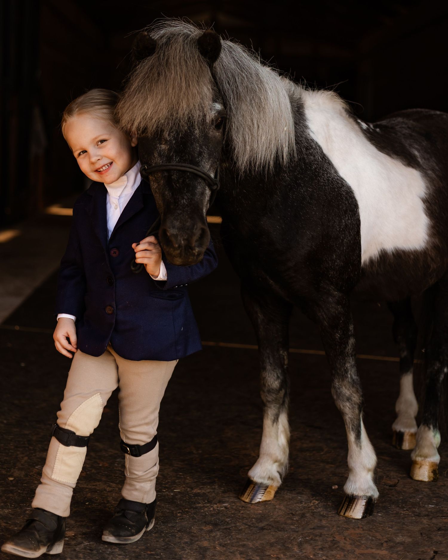 Young child in equestrian attire smiling beside a small black and white pony inside a stable in Oklahoma for equine photoshoot